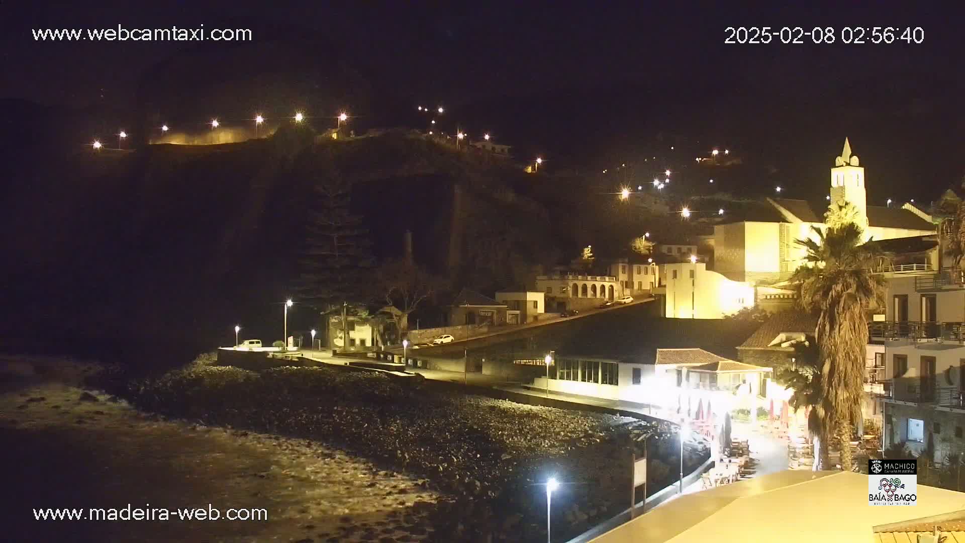 A nighttime view of a coastal town illuminated by streetlights, with a church prominent on a hillside overlooking a dark, rocky beach.