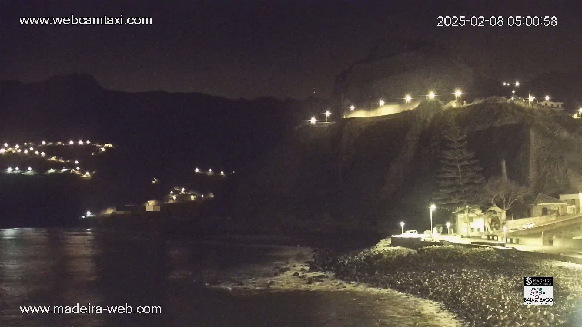 A nighttime coastal scene shows a dark, rocky cliffside with illuminated buildings and roads, along with a calm, dark ocean.