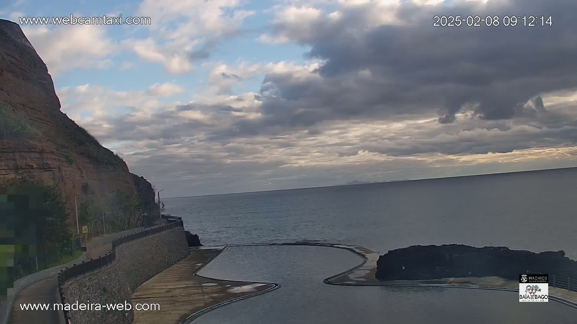 A mostly cloudy sky overlooks a natural rock pool next to a cliffside road, with a calm ocean extending to the horizon.