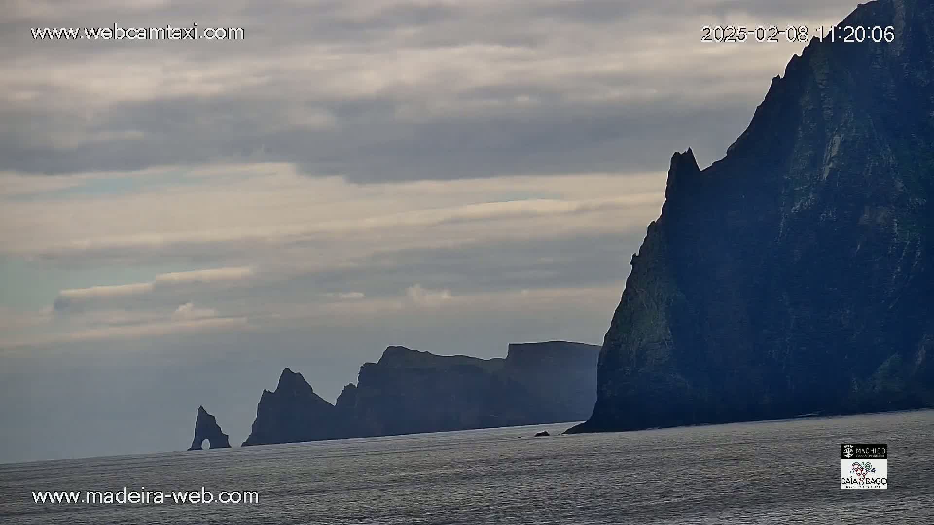 Dark, rocky cliffs rise from a calm sea under a cloudy sky.