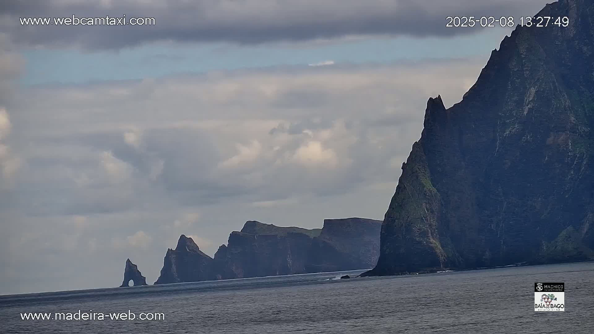 A line of dark, rocky cliffs rises from a calm ocean under a cloudy sky.