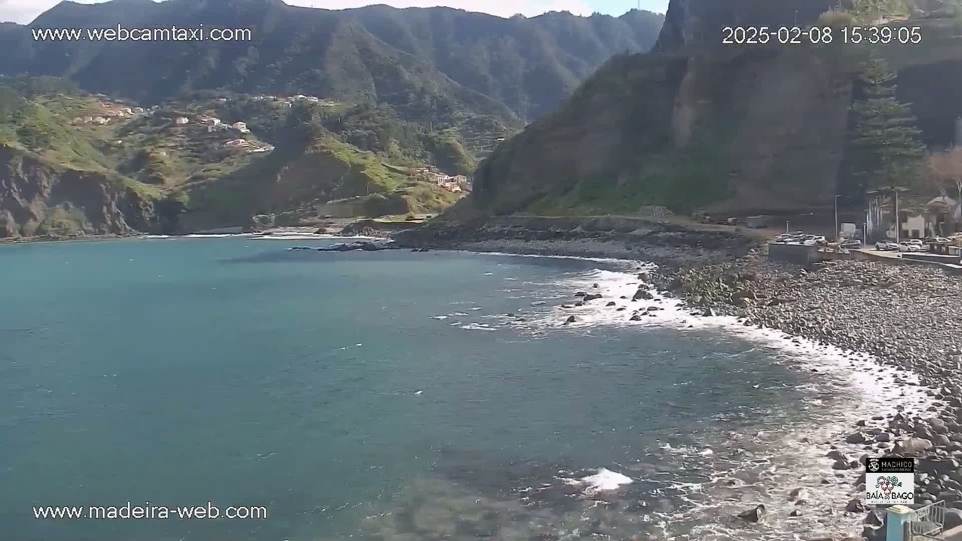 A rocky shoreline with a calm, teal ocean meets a hillside with small buildings under a sunny sky.