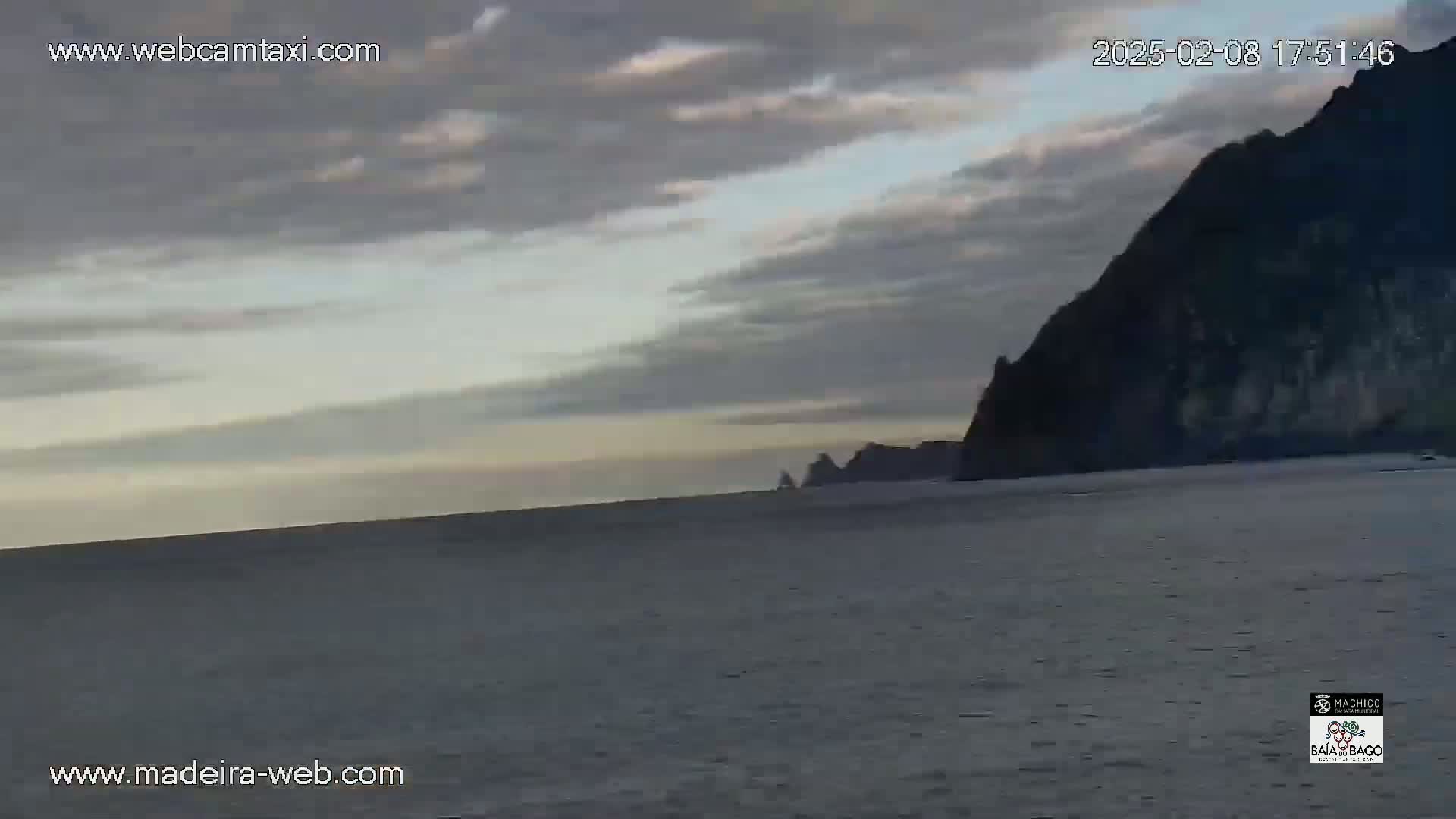 A calm body of water stretches toward a dark, rocky cliff under a mostly cloudy sky.