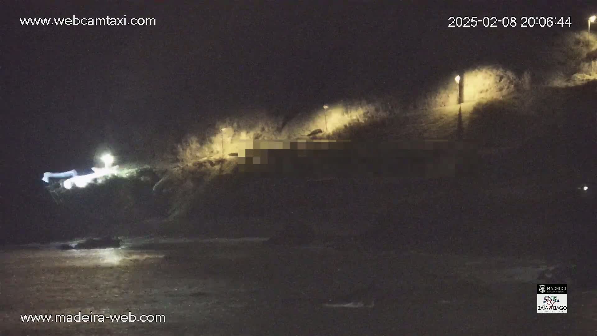 A nighttime coastal scene shows a dark, rocky shoreline illuminated by several lights, with a faintly visible body of water in the foreground.