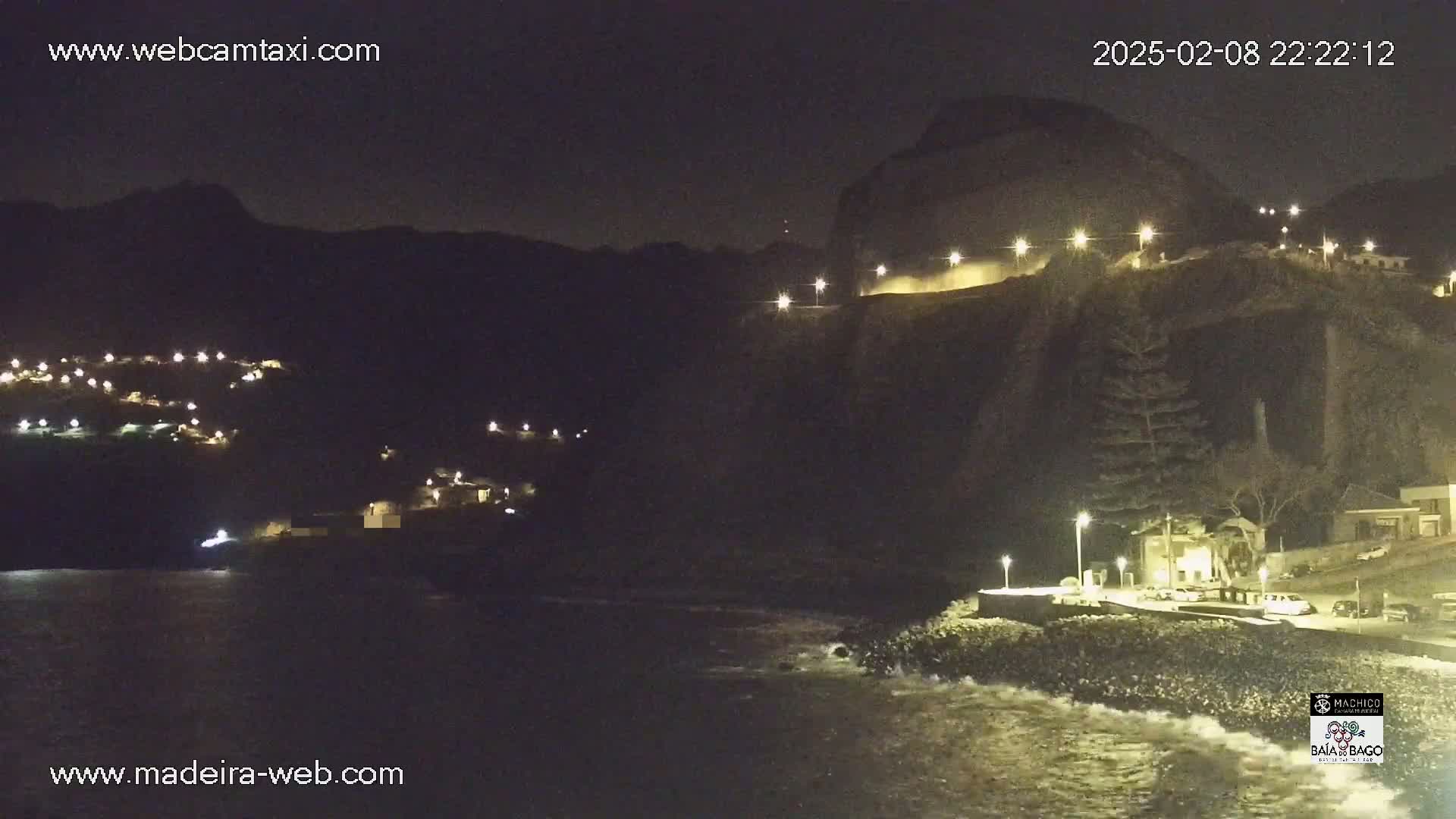 A nighttime coastal scene shows a dark cliff face with lights illuminating a road and buildings, and a dark body of water with small waves lapping the shore.