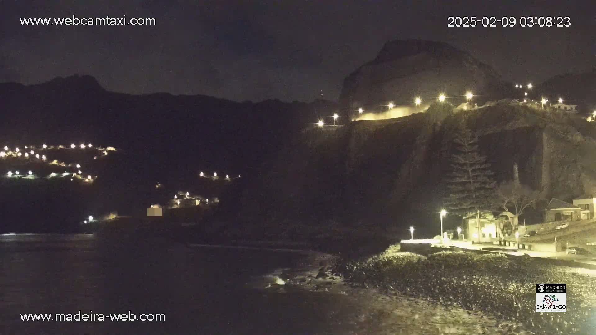 A nighttime coastal scene shows a dark cliff face with lights illuminating a road along its top and a small town nestled on a hillside below, with a dark ocean at the bottom.
