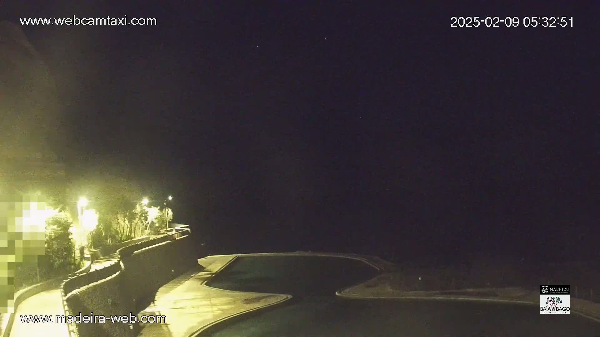 A nighttime view of a winding road and wall next to a dark body of water, illuminated by streetlights under a clear, starlit sky.