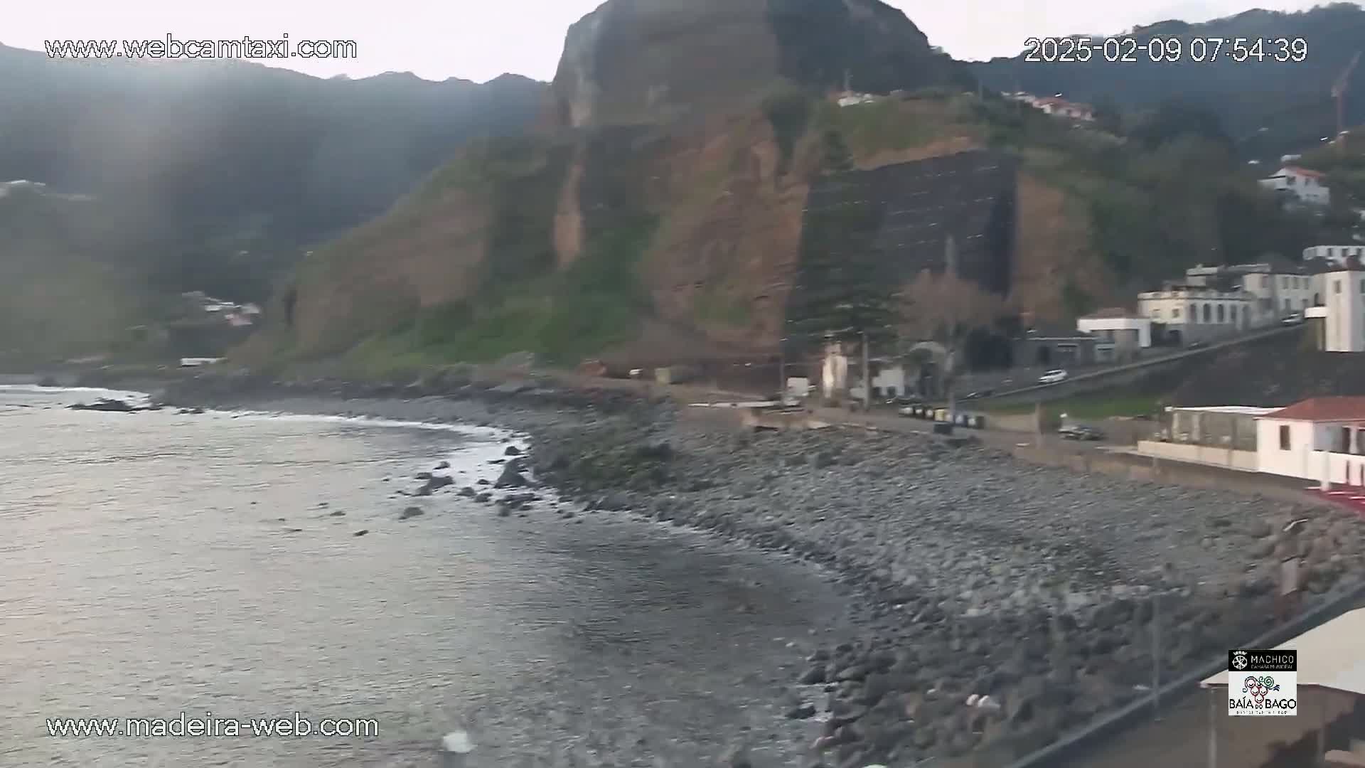 A rocky shoreline with calm water meets a hillside town under overcast skies.