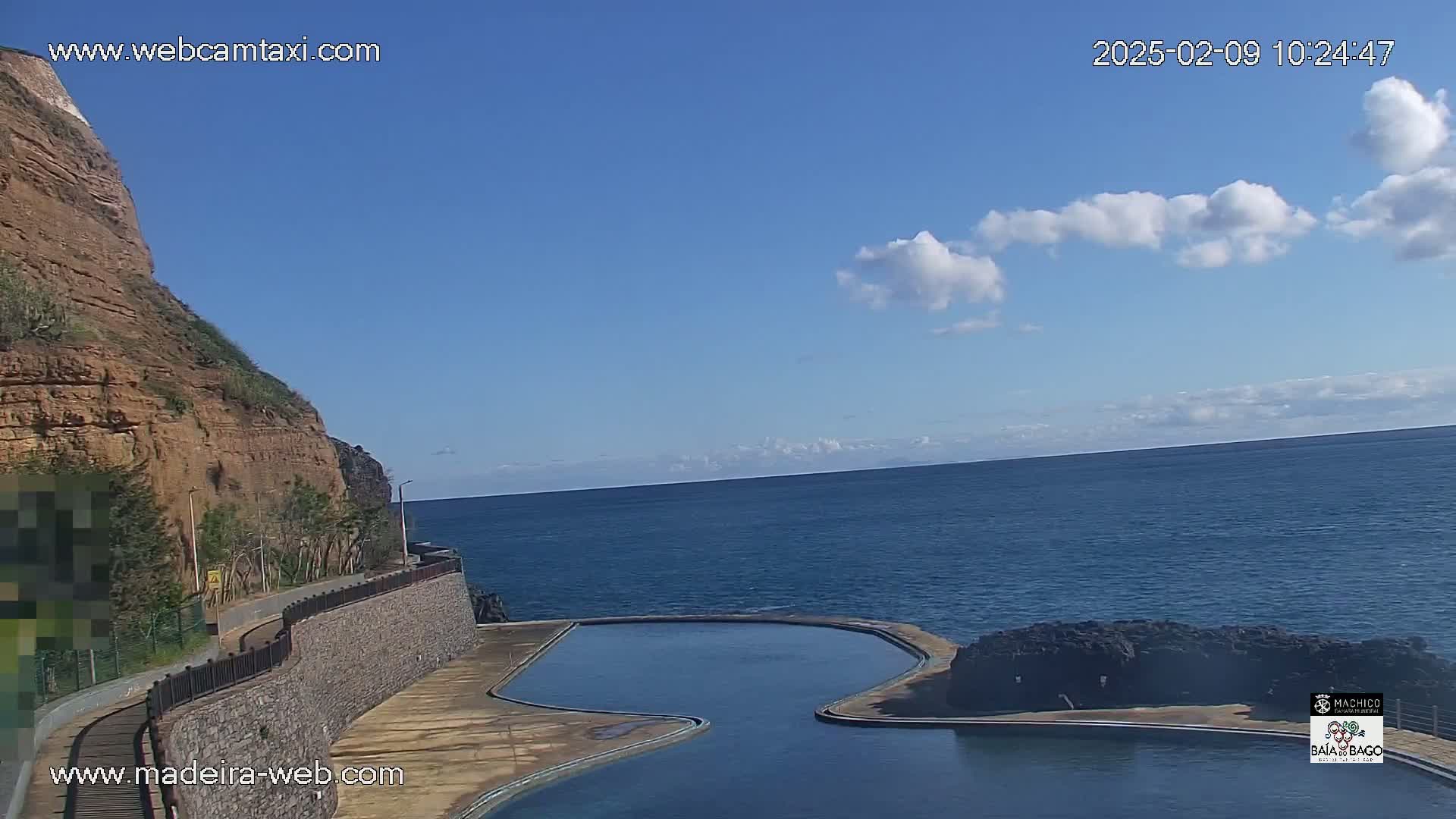 A seaside scene under a mostly clear blue sky with a few clouds shows a rocky cliff overlooking a calm ocean and a curving, man-made pool.