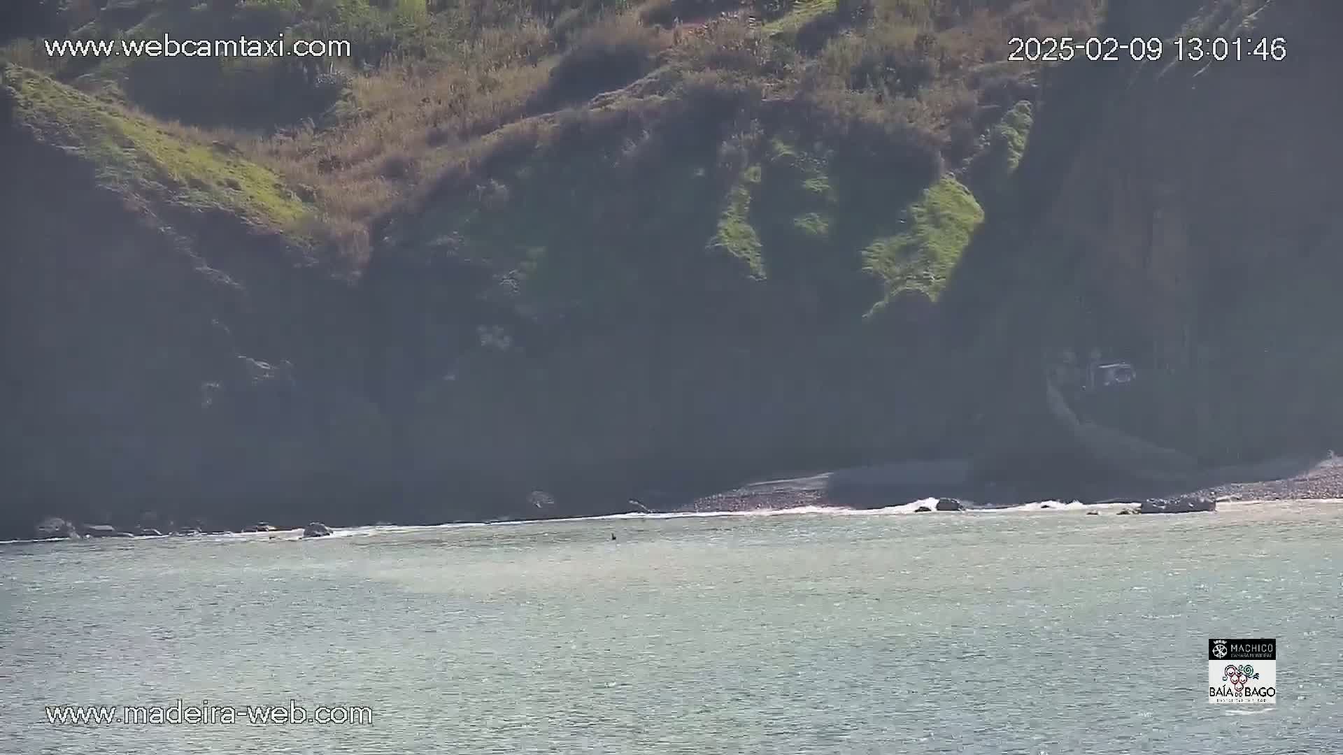 A rocky, vegetated cliff overlooks a calm ocean with small waves lapping a pebble beach.
