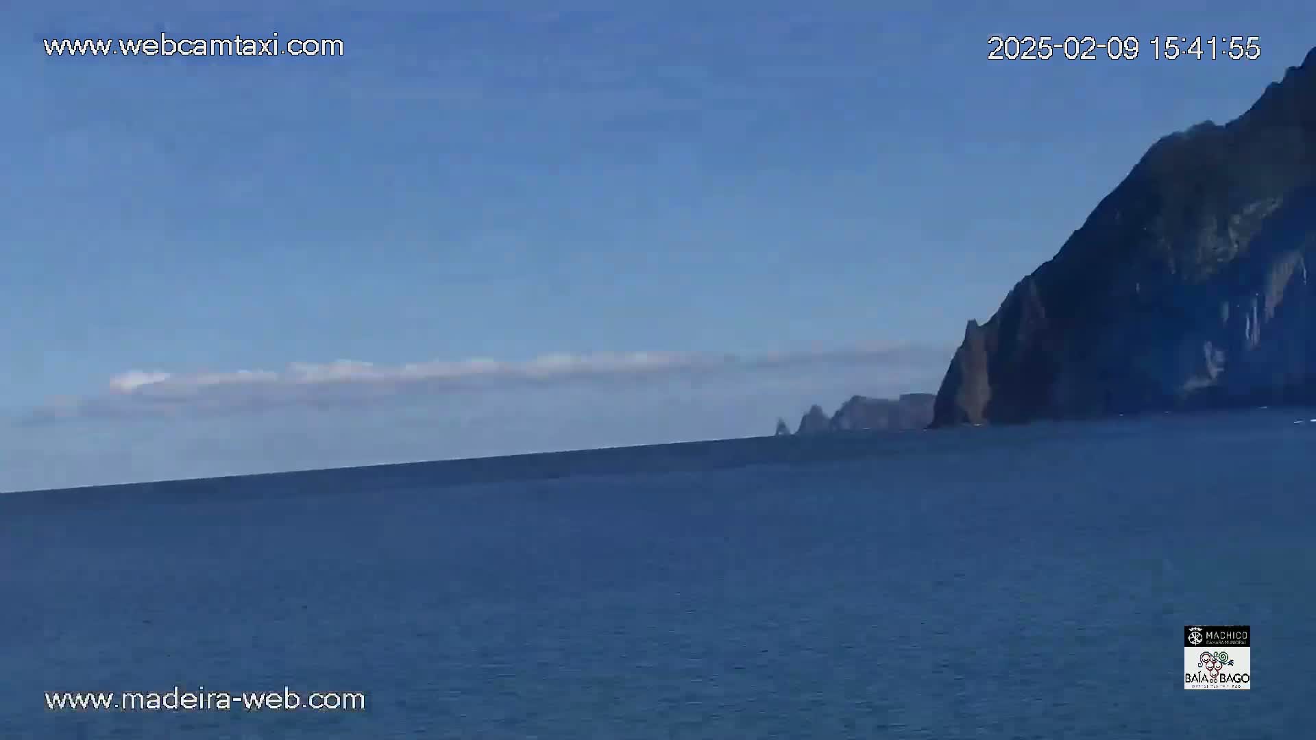 A calm ocean stretches to a distant, dark cliff face under a mostly clear sky with a thin layer of clouds.