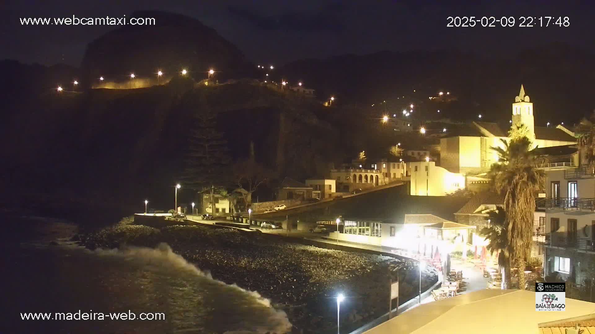 A nighttime coastal town scene with a rocky beach and crashing waves, illuminated by streetlights, showing buildings nestled against a dark hillside.