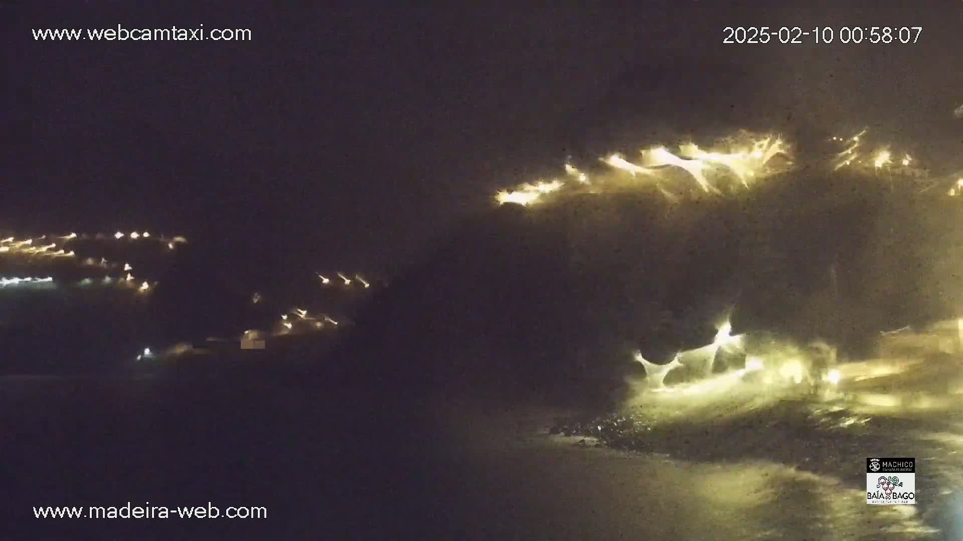 A nighttime coastal scene shows waves crashing against a dark shore illuminated by lights from a town in the distance, under a dark, stormy sky.