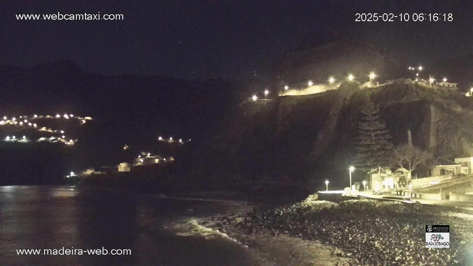 A nighttime coastal scene shows a dark, rocky cliff with lights illuminating a road and buildings along its edge, while a village is nestled on a hillside across a dark body of water.