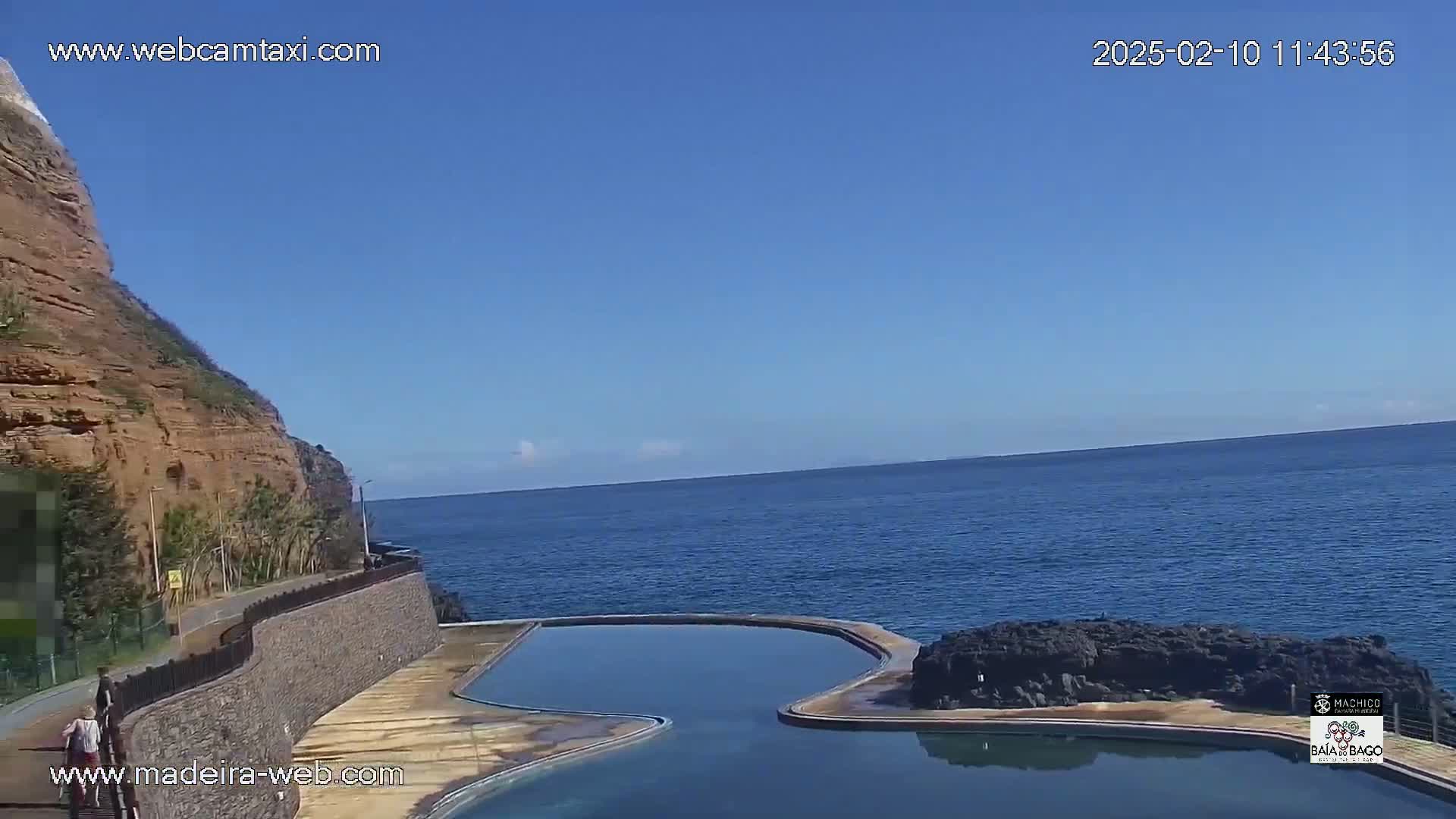 An outdoor scene shows a swimming pool next to a rocky cliff overlooking a calm ocean under a clear blue sky.