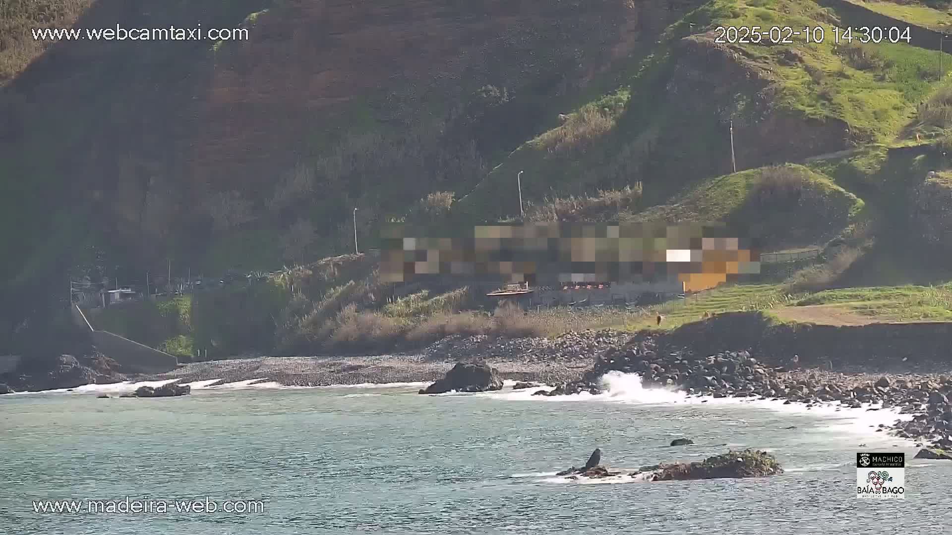 A rocky beach with small waves laps against the shore below a grassy hillside on a sunny day.