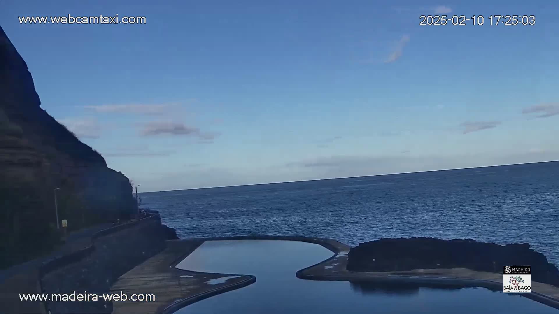 A calm ocean stretches to the horizon under a mostly clear blue sky, beside a dark cliff and a man-made pool.
