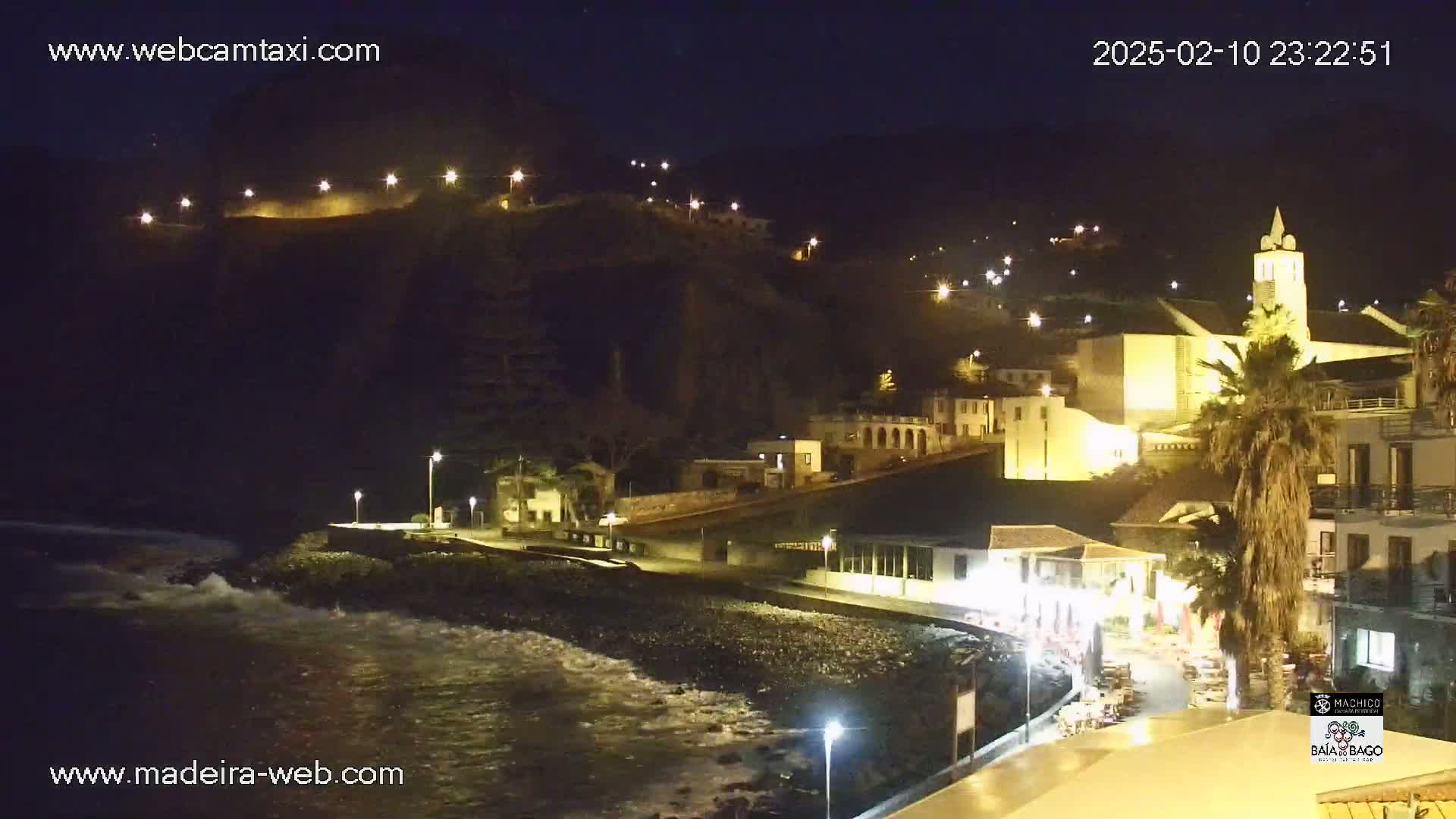 A nighttime coastal town with illuminated buildings and a church, overlooking a rocky shore with gentle waves under a clear, dark sky.