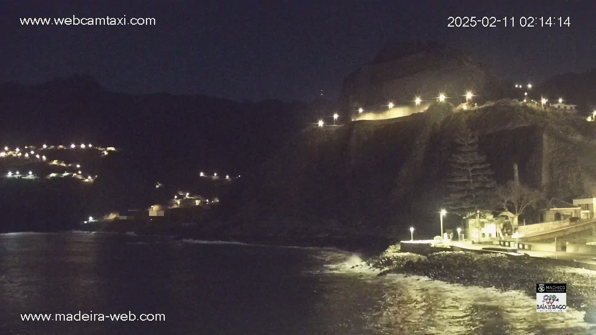 A nighttime coastal scene shows a dark cliffside with buildings illuminated by lights, overlooking a dark ocean with small waves.