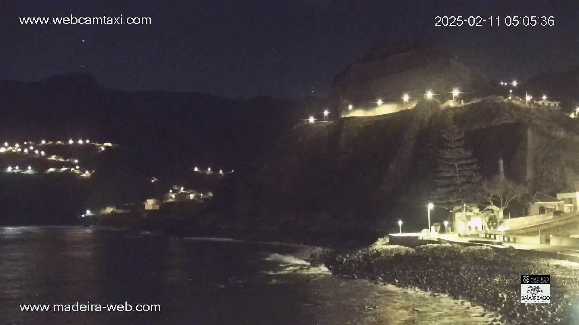 A nighttime coastal scene shows a dark cliff with lights illuminating a roadway and buildings along its edge, overlooking a dark ocean with gentle waves.