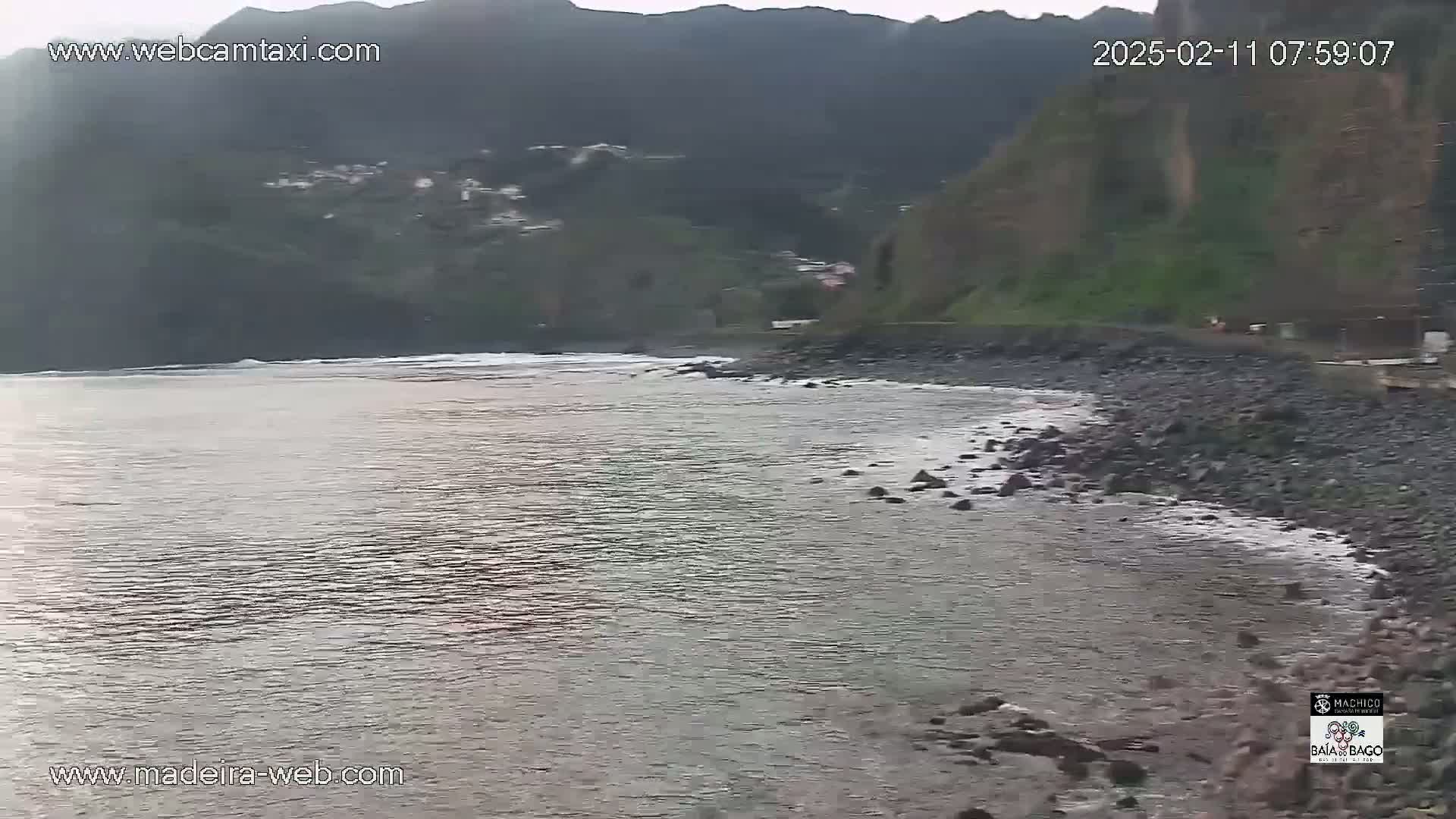 A rocky shoreline meets a calm, gray ocean under an overcast sky, with dark green hills rising in the background.