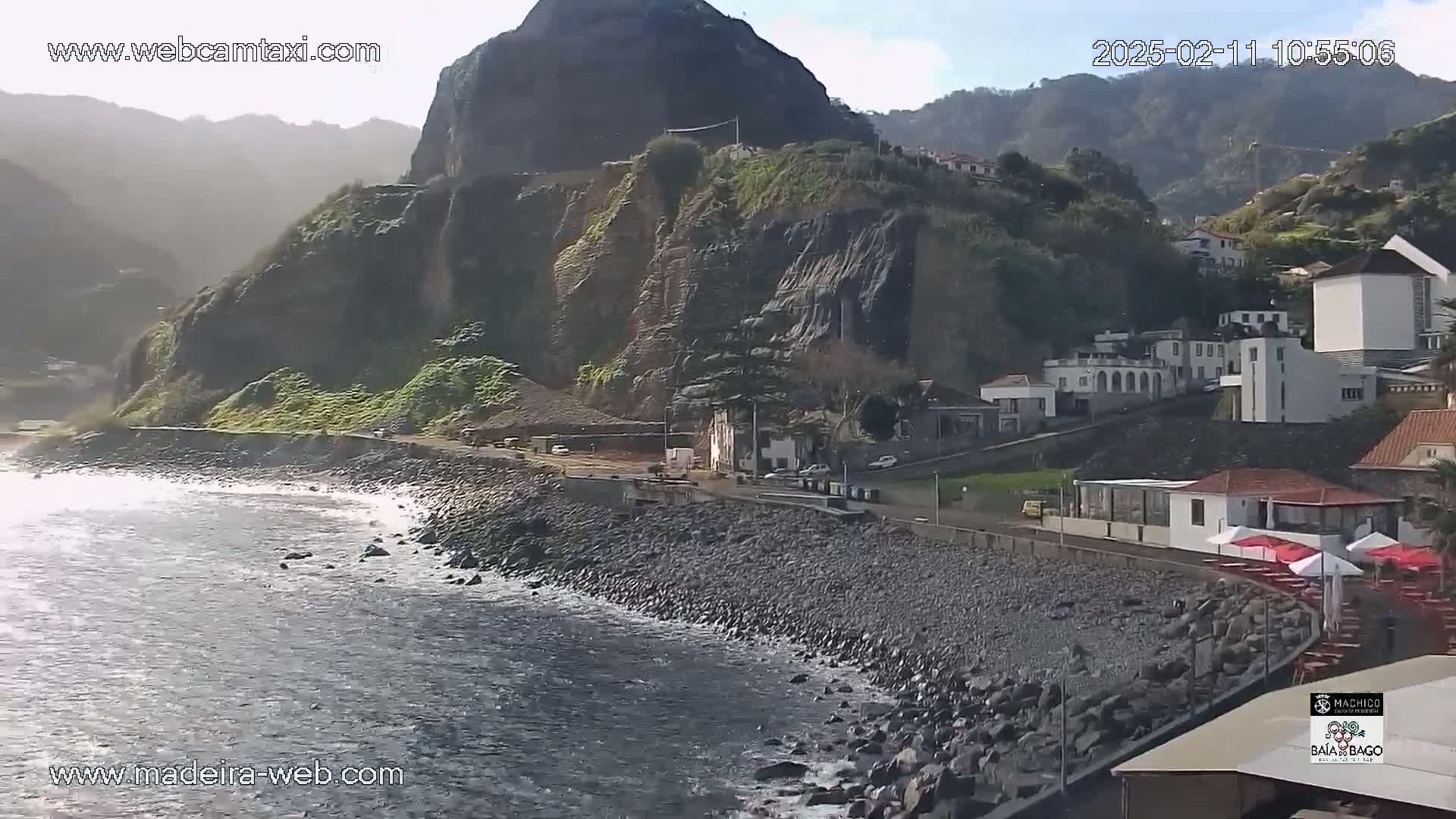 A rocky coastline with a small village nestled against a steep, dark cliff faces a calm ocean under a mostly sunny sky.