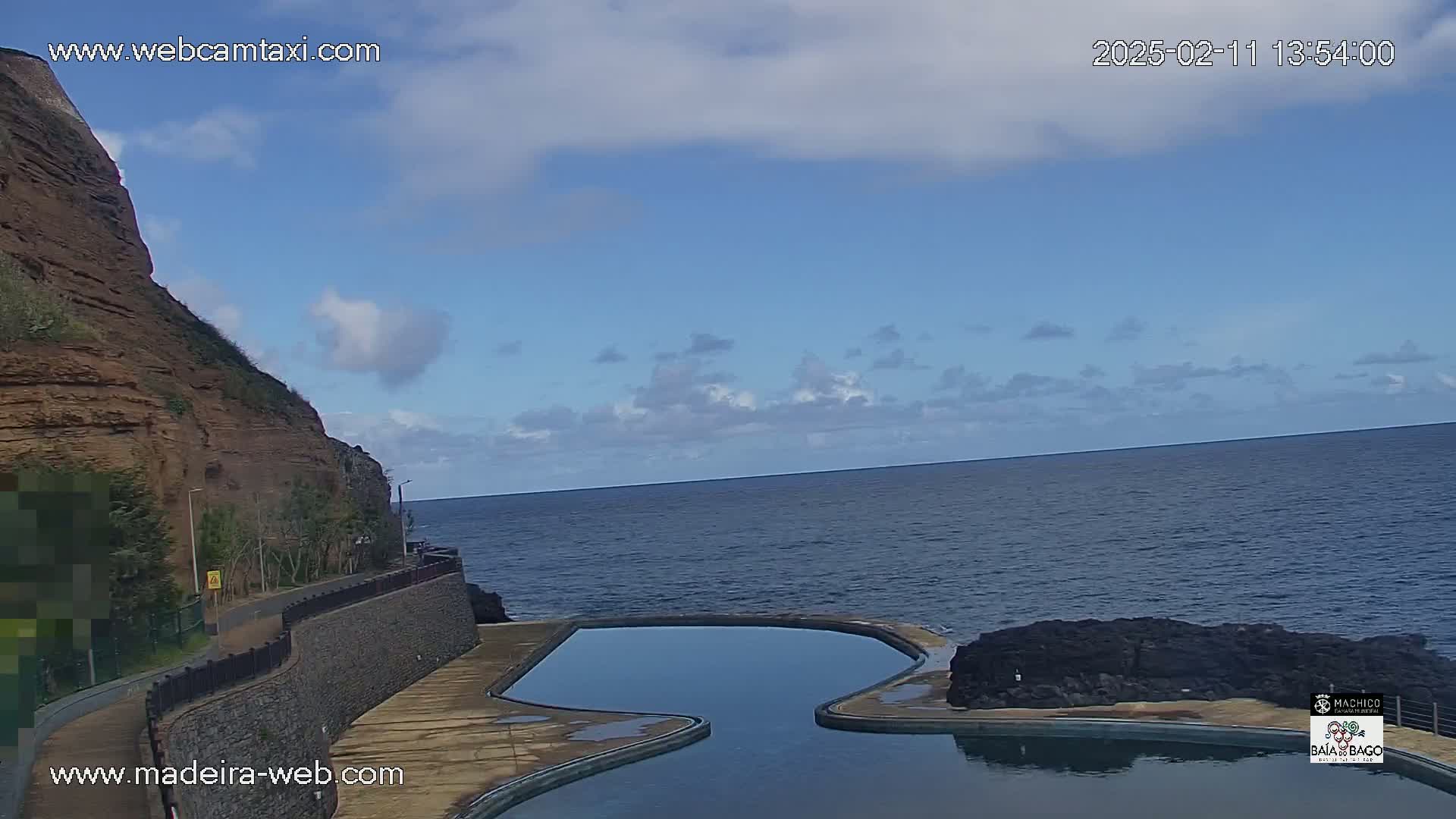 A partly cloudy day shows a rocky cliff overlooking a dark blue ocean and an irregularly shaped swimming pool next to the water.