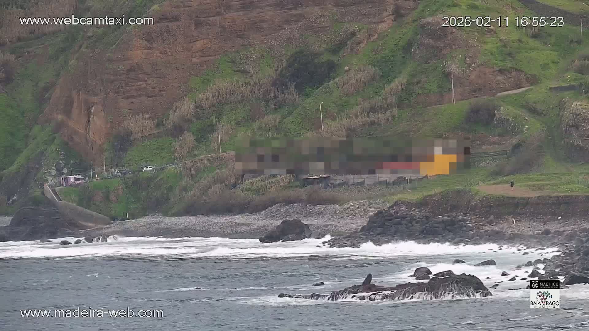 A rocky coastline with a small building near a pebble beach, with moderate waves breaking on the shore under overcast skies.
