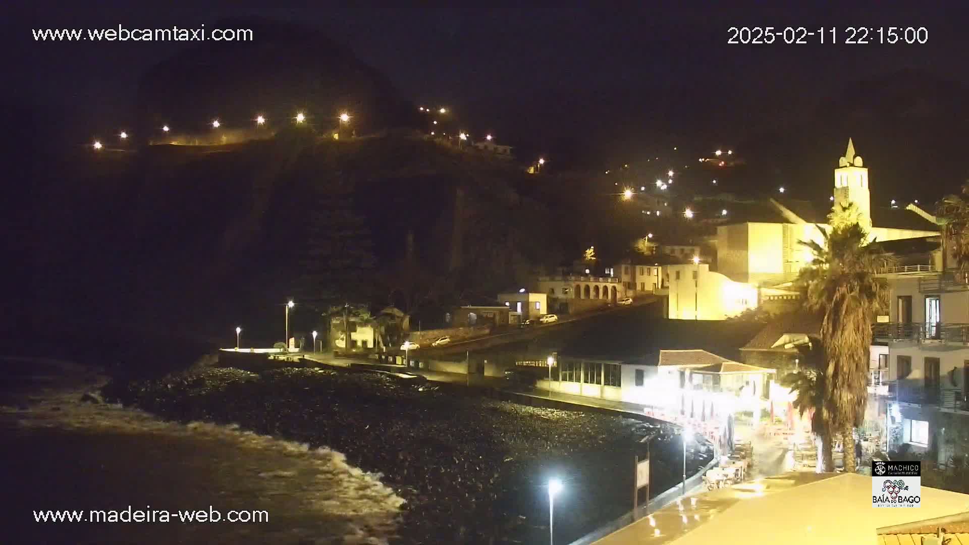 A nighttime coastal town is illuminated by streetlights, with waves crashing on a dark beach in the foreground.
