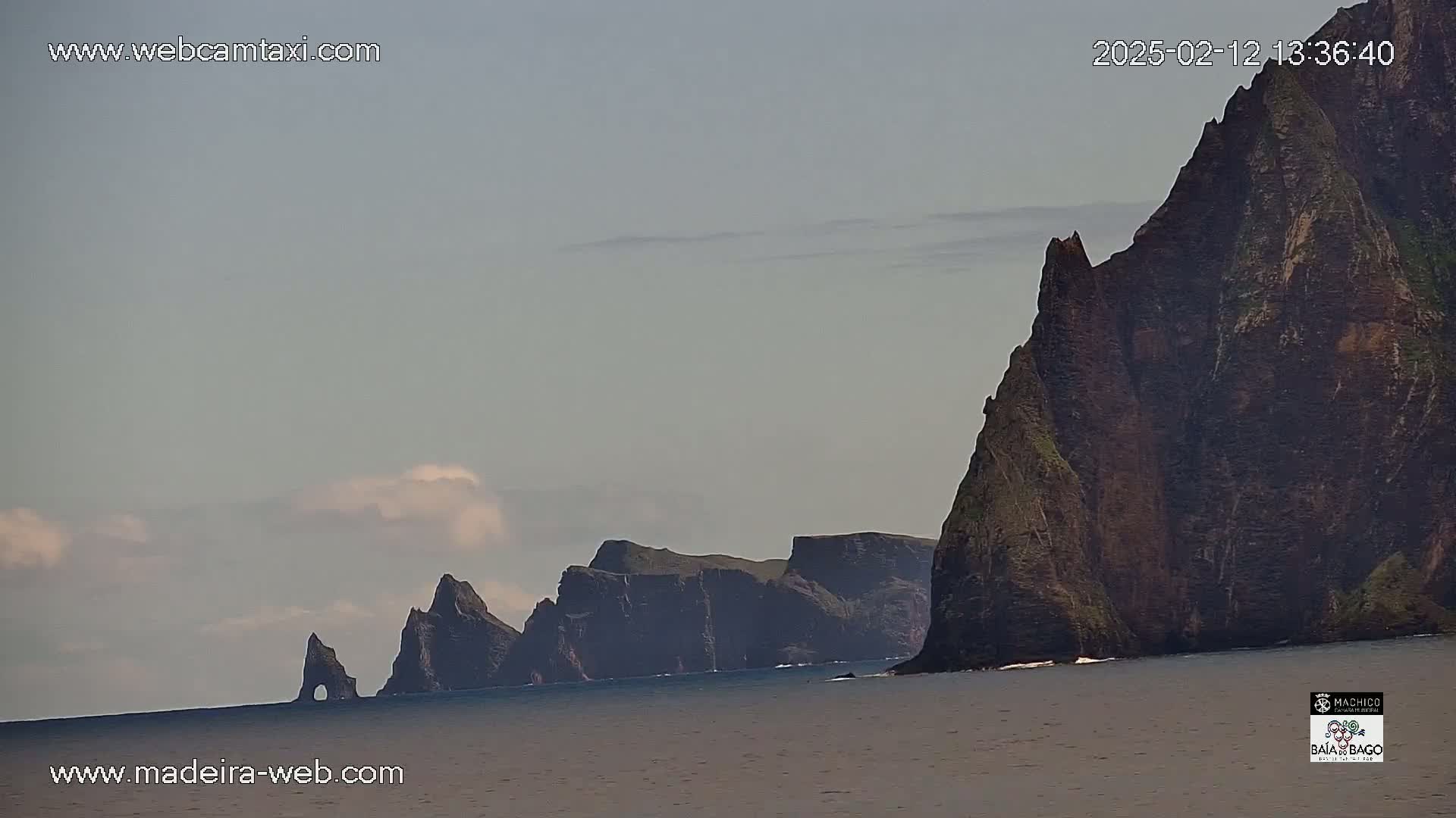 A mostly cloudy sky overlooks dark, rocky cliffs and sea stacks jutting from the ocean.