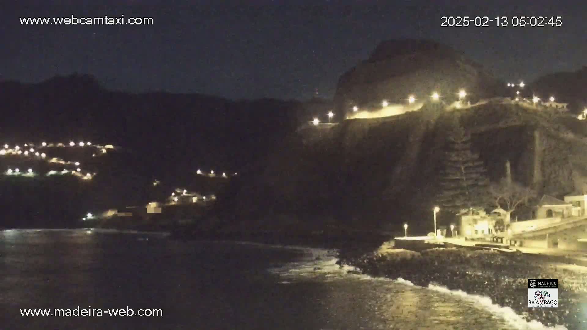 A nighttime coastal scene shows a dark, rocky cliff illuminated by lights, with a town nestled below on a hillside and gentle waves lapping at the shore.