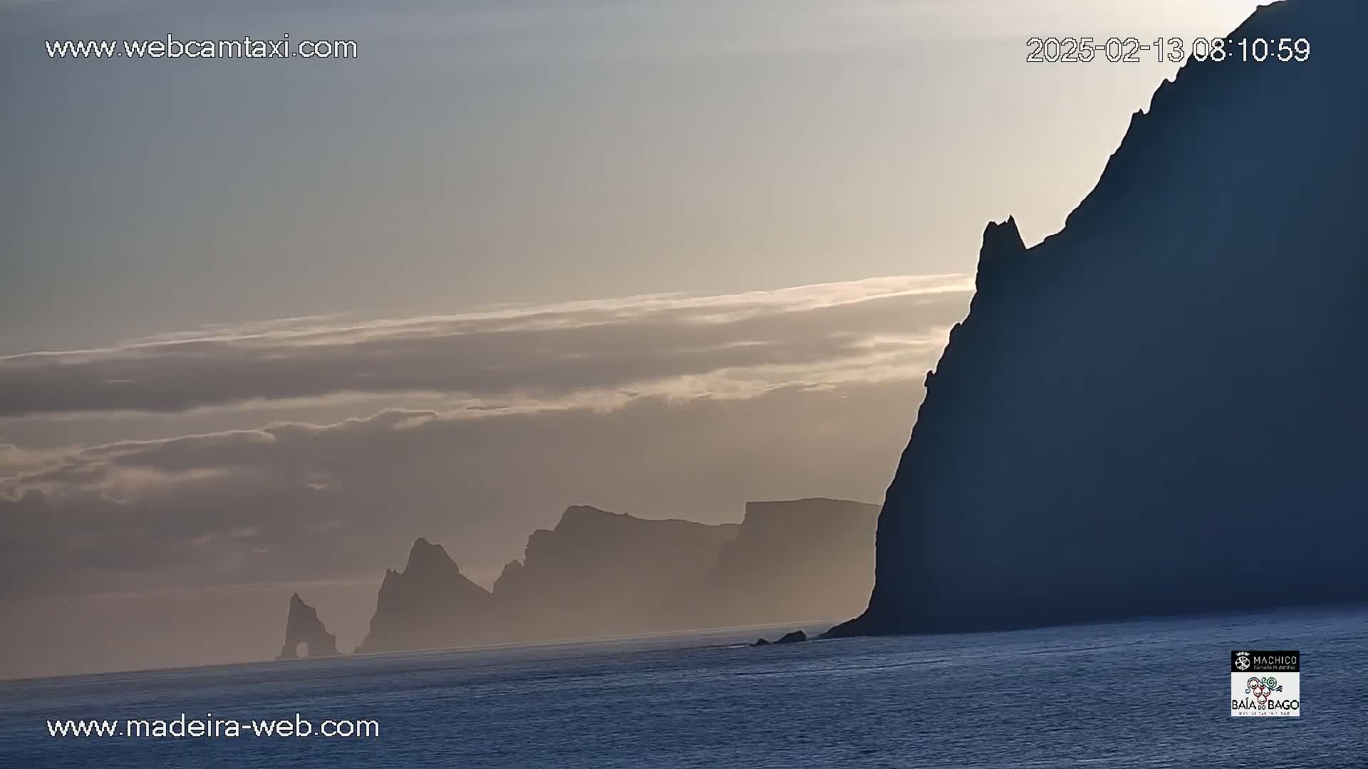 Silhouetted cliffs and rock formations rise from a calm ocean under a partly cloudy, pale sky.