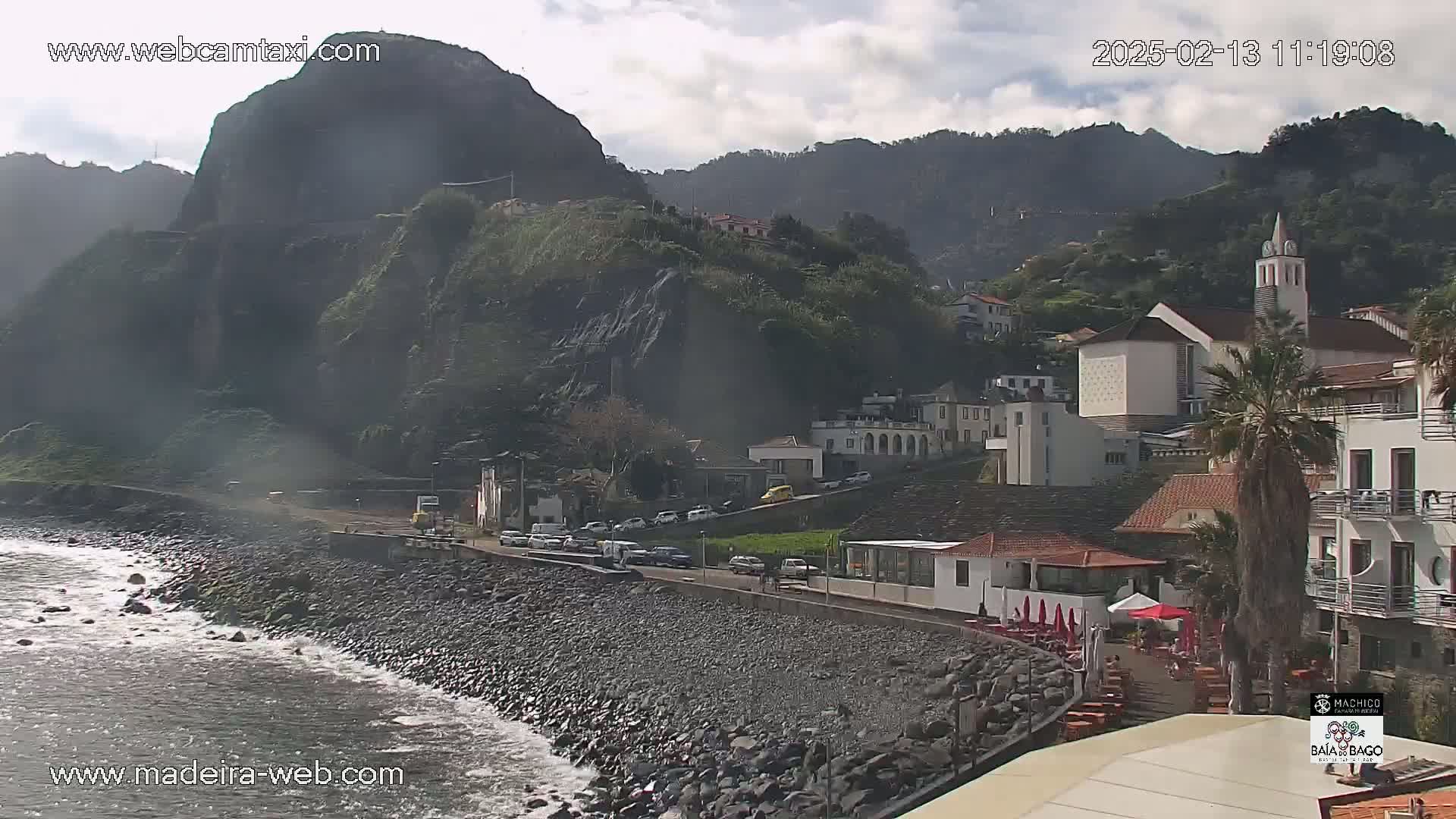 A rocky coastline with a village nestled against steep, dark green mountains under a partly cloudy sky.