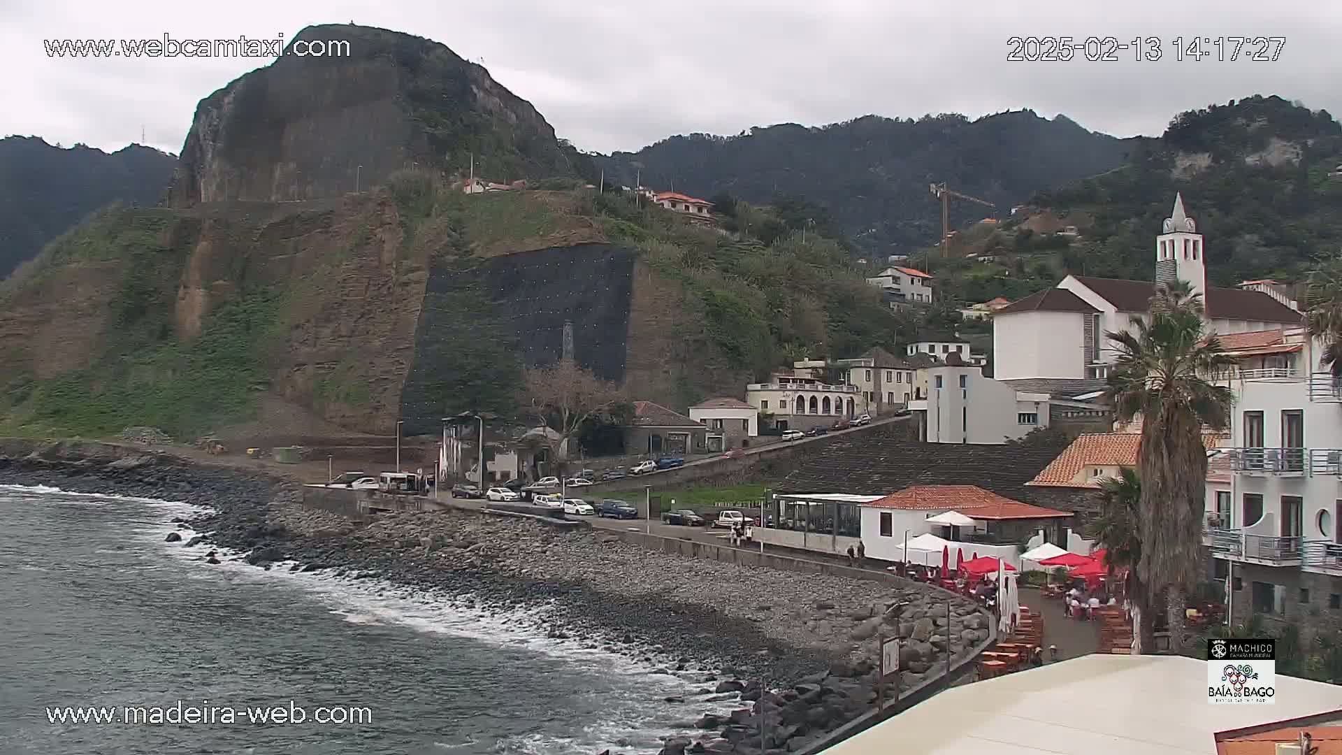 A rocky coastline with a village nestled against a hillside under an overcast sky, featuring a church and several buildings along the shore.