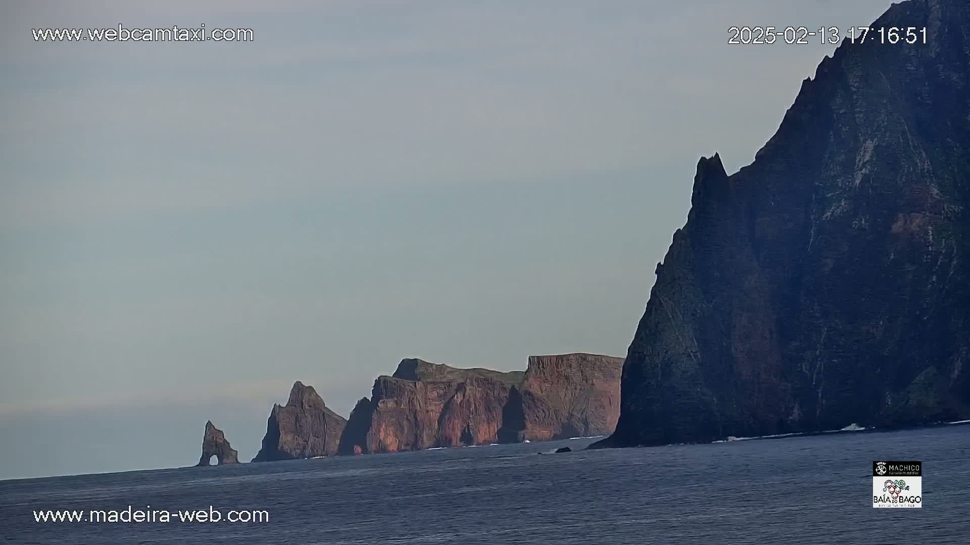 A line of dark, rocky cliffs extends across a calm ocean under a pale, overcast sky.