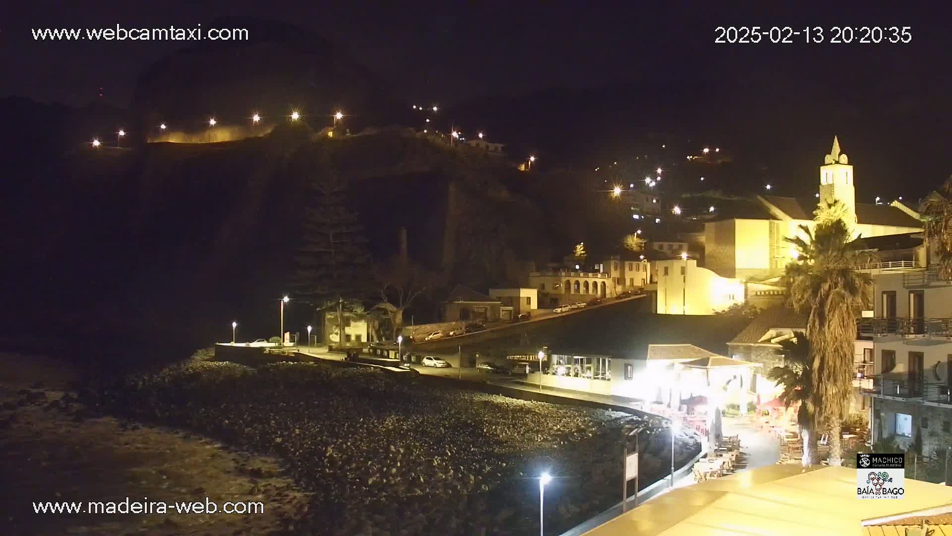 A nighttime view of a coastal town, illuminated by streetlights, nestled against a dark hillside with a church prominent in the center.