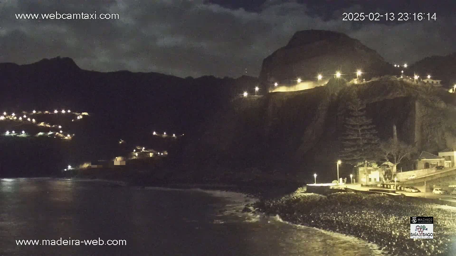 A nighttime coastal scene shows a dark, rocky cliff face with lights illuminating a road along the top and buildings below, while gentle waves lap at the shore under a partly cloudy sky.