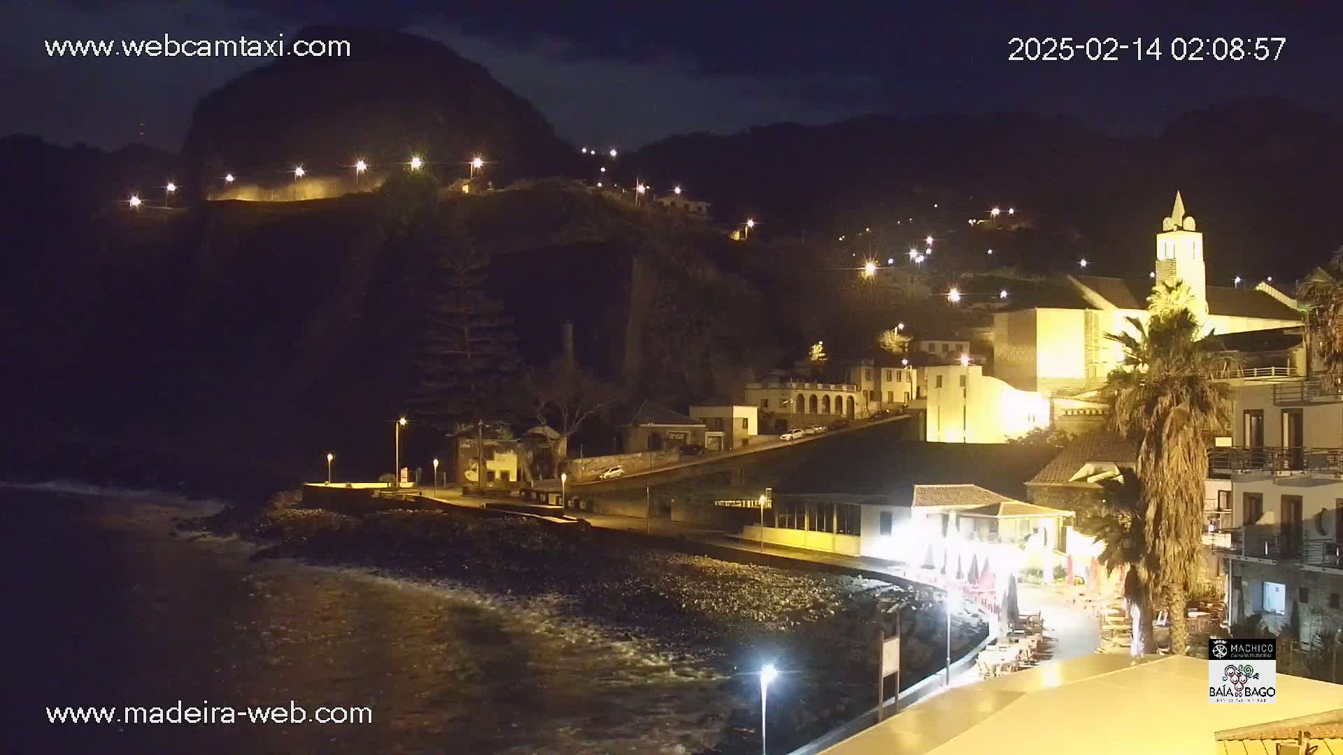 A nighttime coastal town nestled against a dark hill, illuminated by streetlights, with gentle waves lapping at a rocky shore.