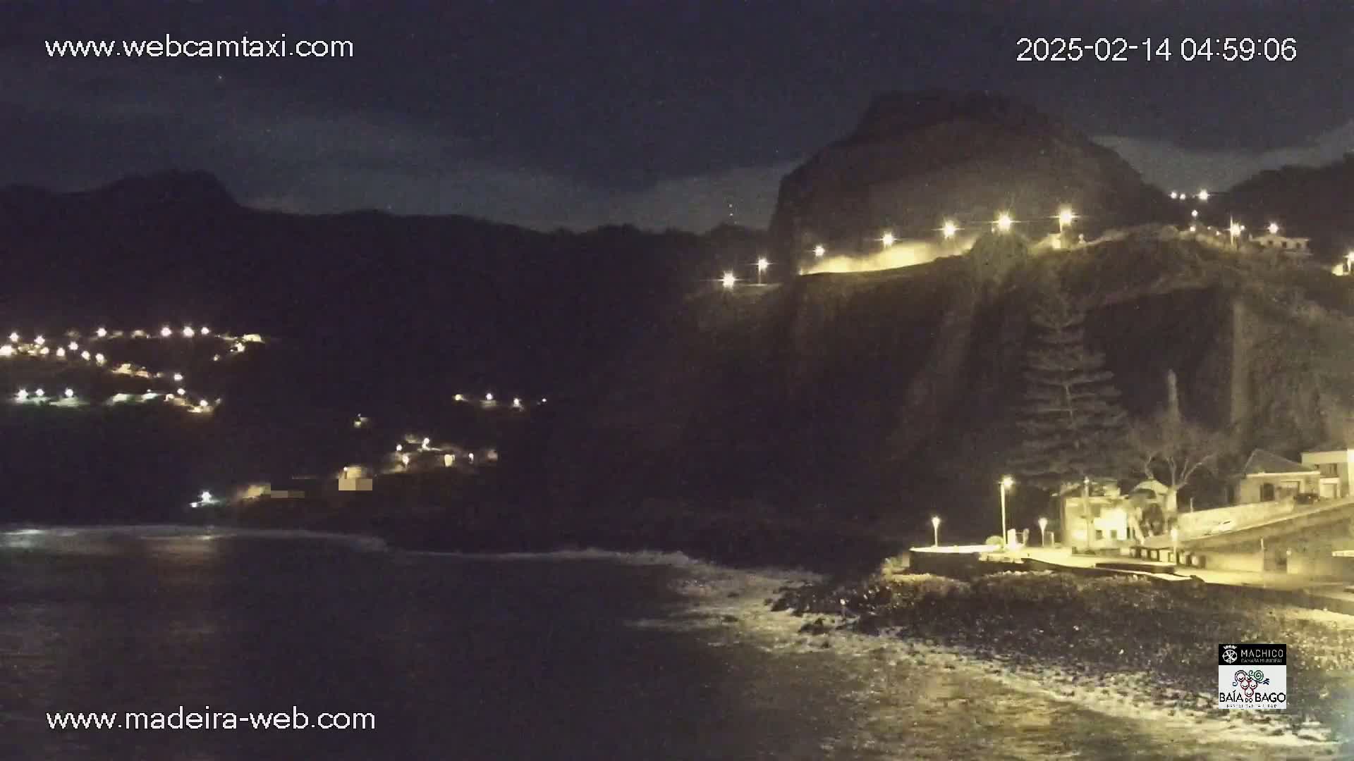 A nighttime coastal scene shows a dark cliff with lights along its top and a village below, illuminated by many small lights, with dark, gently rolling ocean waves at the bottom.