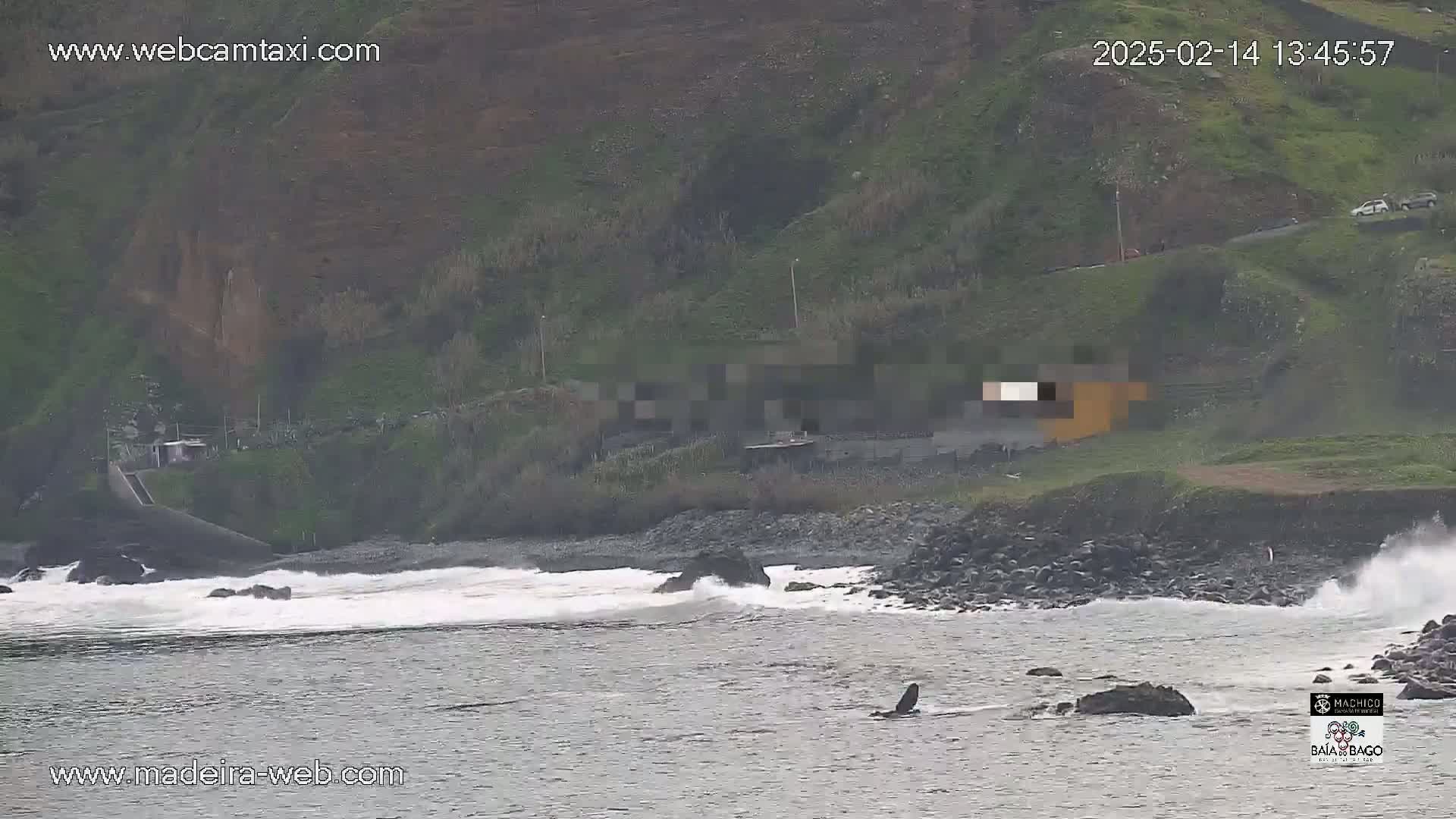 Waves crash on a rocky shore near a hillside with some buildings, under an overcast sky.