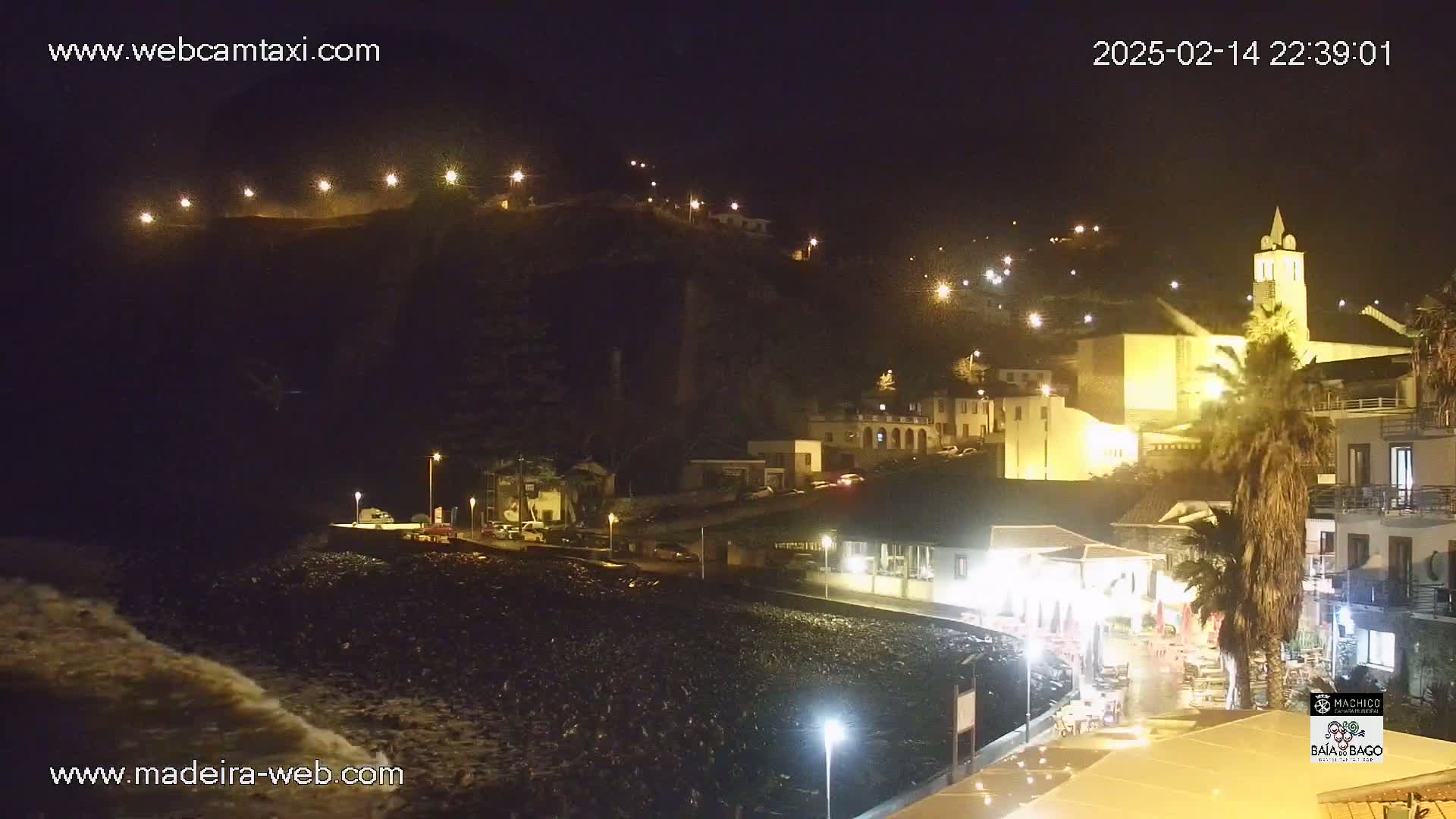 A nighttime coastal town illuminated by streetlights, with a dark, rocky beach and waves crashing ashore.