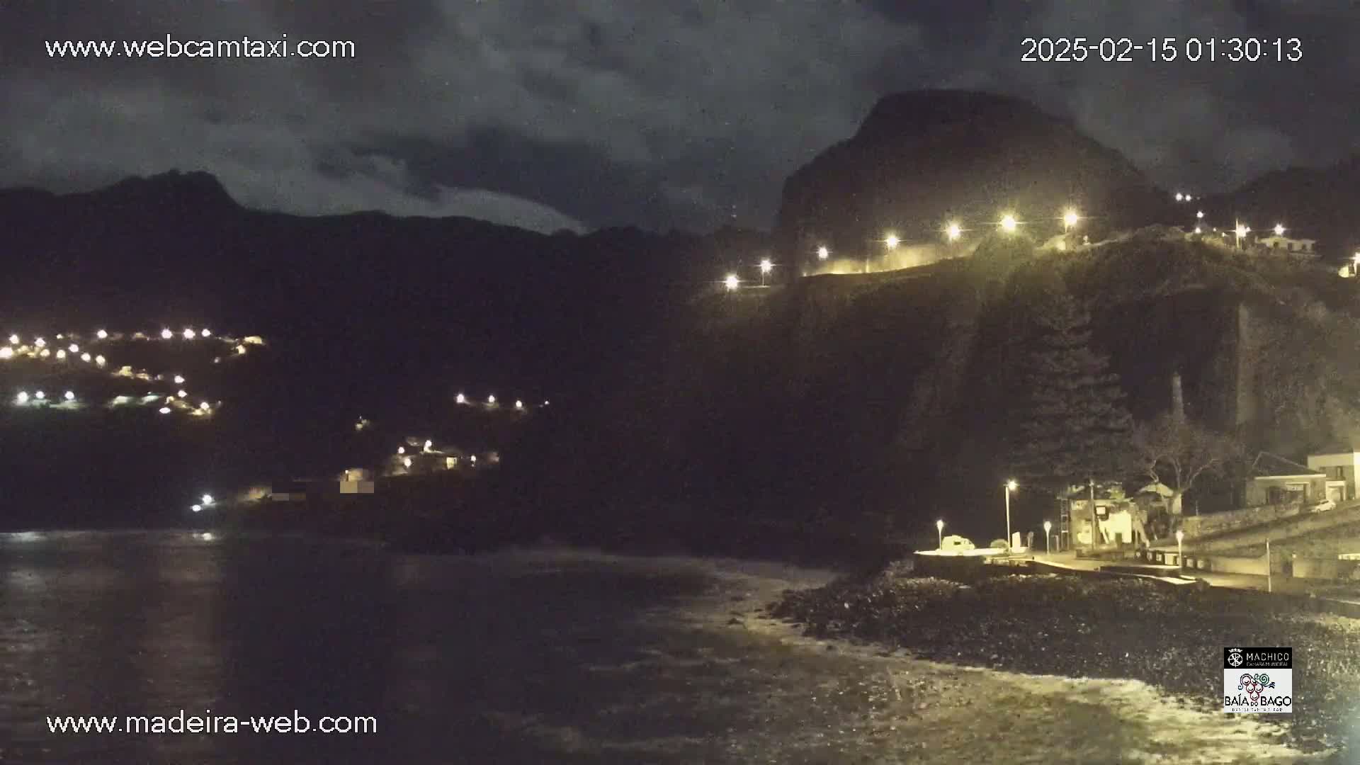 A nighttime coastal scene shows a dark, mountainous landscape with illuminated buildings and roads, overlooking a dark ocean with gentle waves under a cloudy sky.