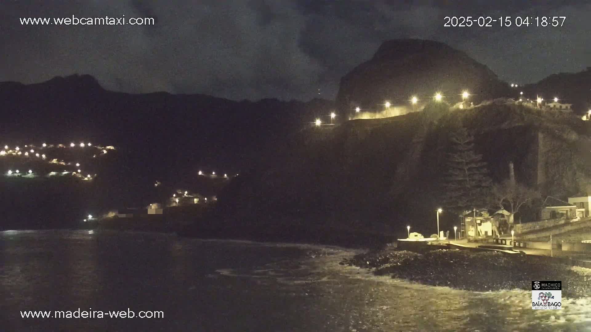 A nighttime coastal scene shows a dark, rocky cliff face with scattered lights along its top and bottom, overlooking a body of water with small waves.
