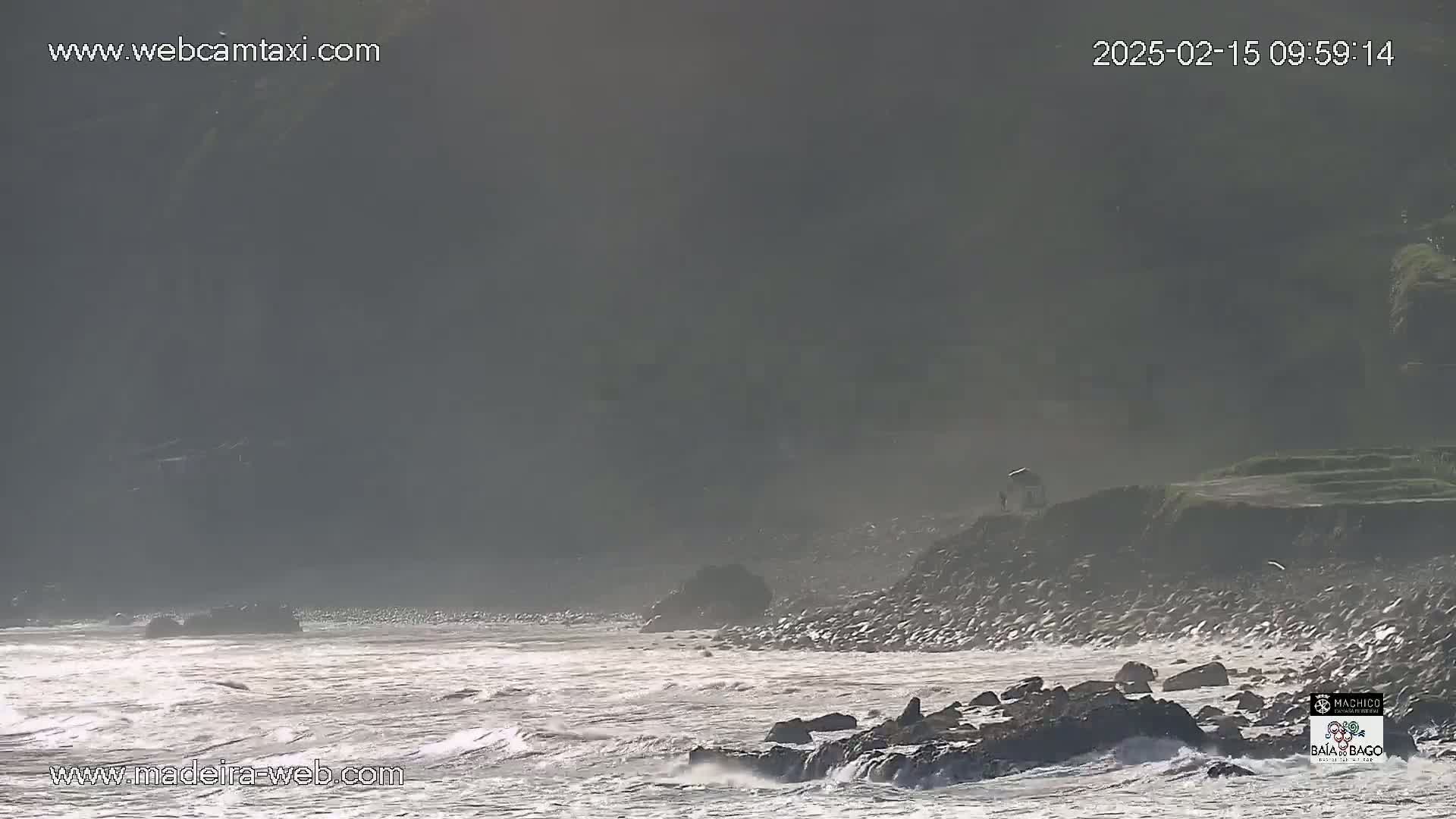 A rocky coastline with rough, gray-white waves under hazy sunlight.