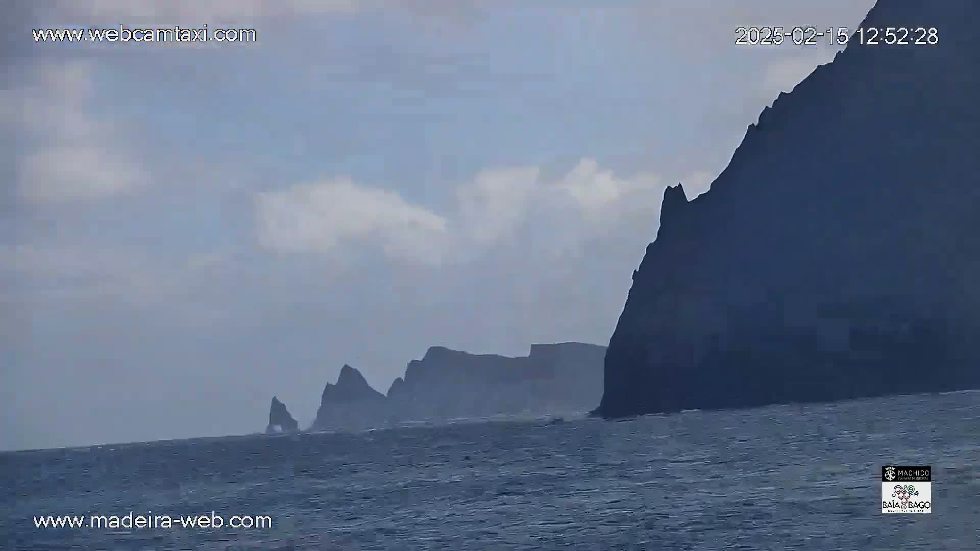 Dark, rocky cliffs rise from a calm, gray ocean under a partly cloudy sky.