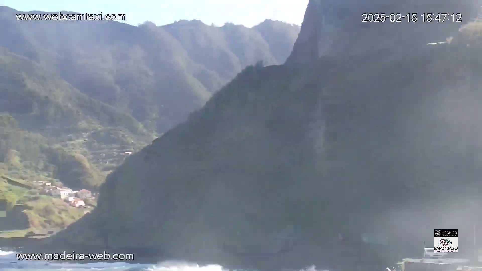 A coastal village nestled in a valley between steep, hazy mountains overlooks a wavy ocean under a clear sky.