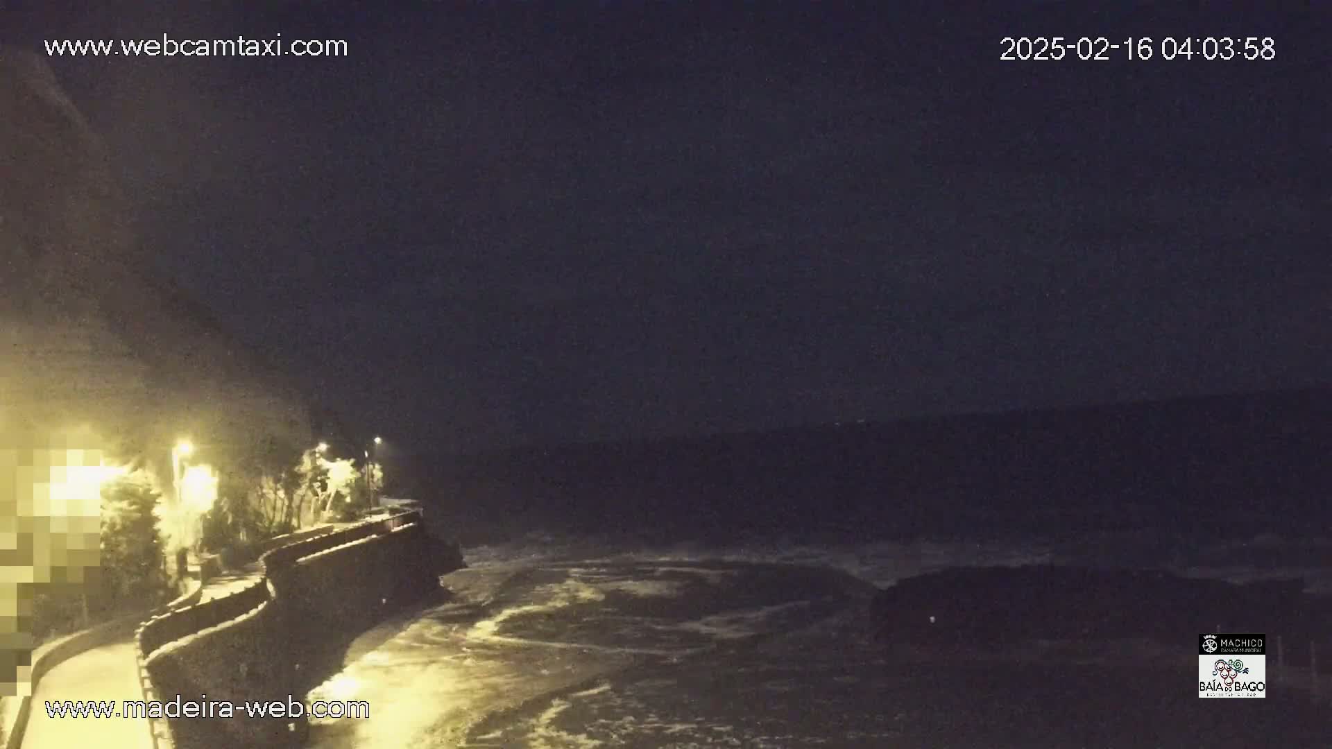 A nighttime coastal scene shows dark, wavy ocean water lapping against a stone wall bordering a walkway lit by streetlights.