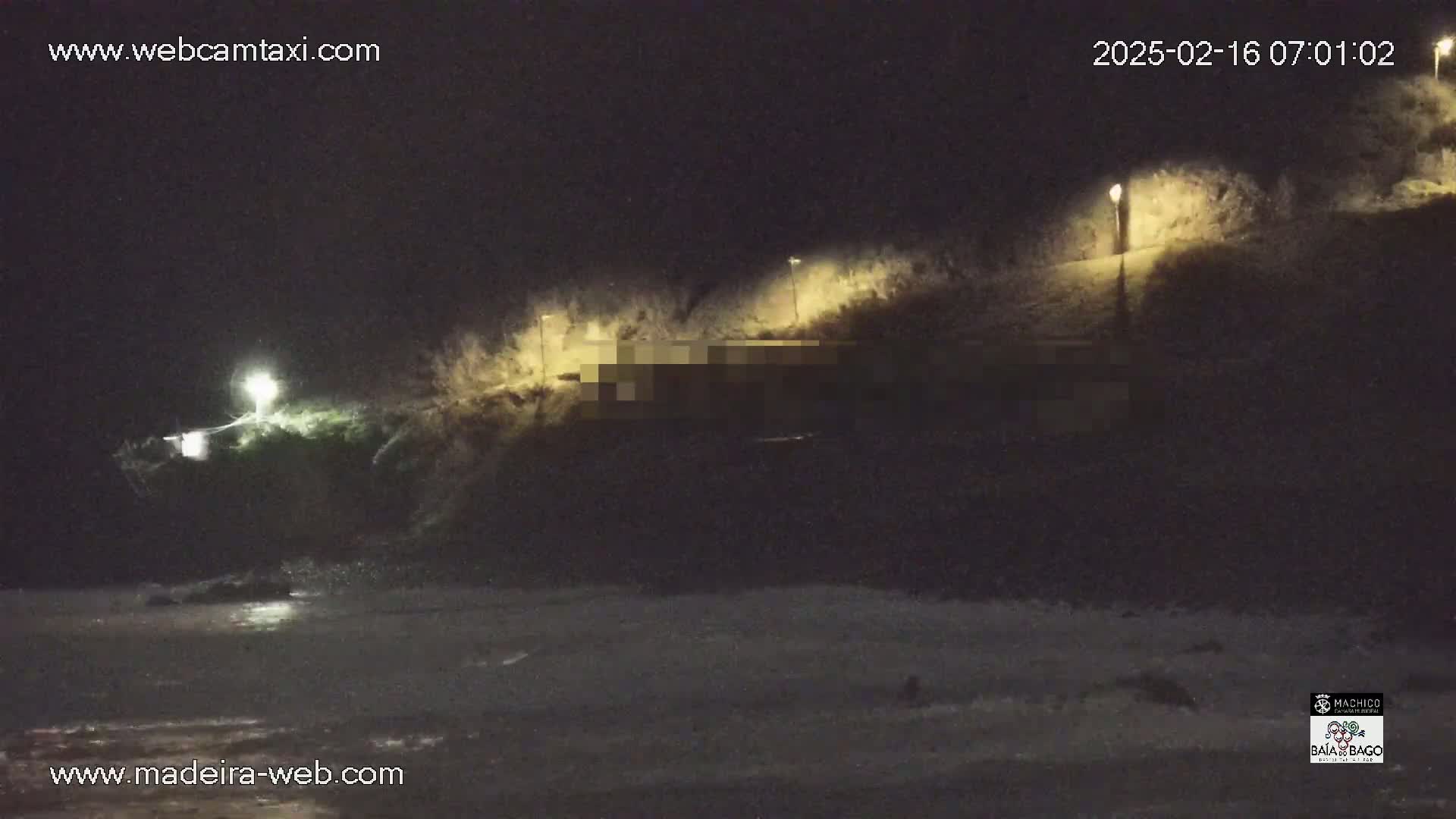 A nighttime coastal scene shows a dark, sloped area with lights illuminating vegetation and a structure, overlooking a dark beach with waves lapping at the shore.