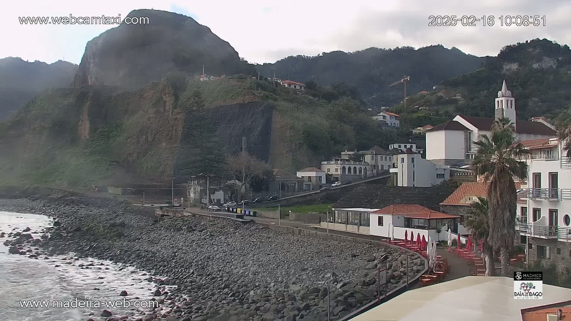 A rocky coastline with gentle waves laps against a pebble beach in front of a hillside village featuring white buildings and a church under overcast skies.