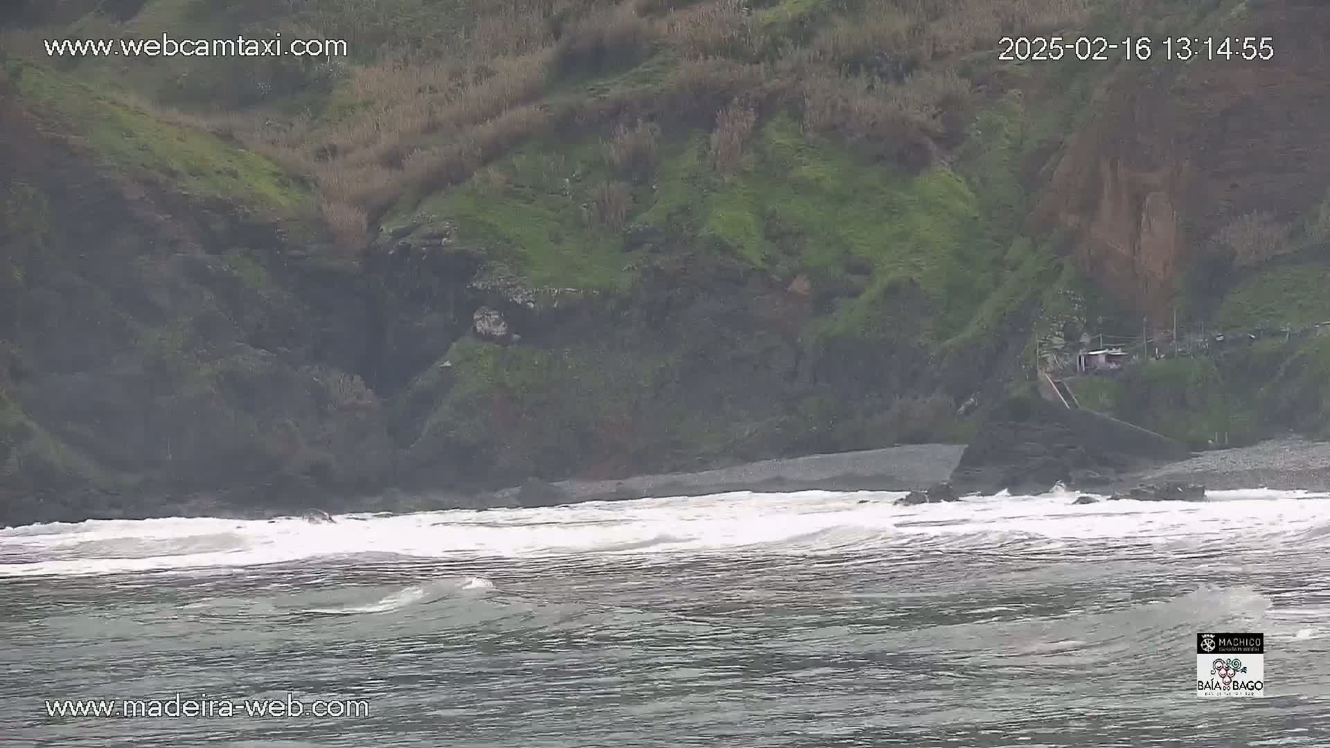 Waves crash on a rocky beach below a green, cliffside hillside on an overcast day.